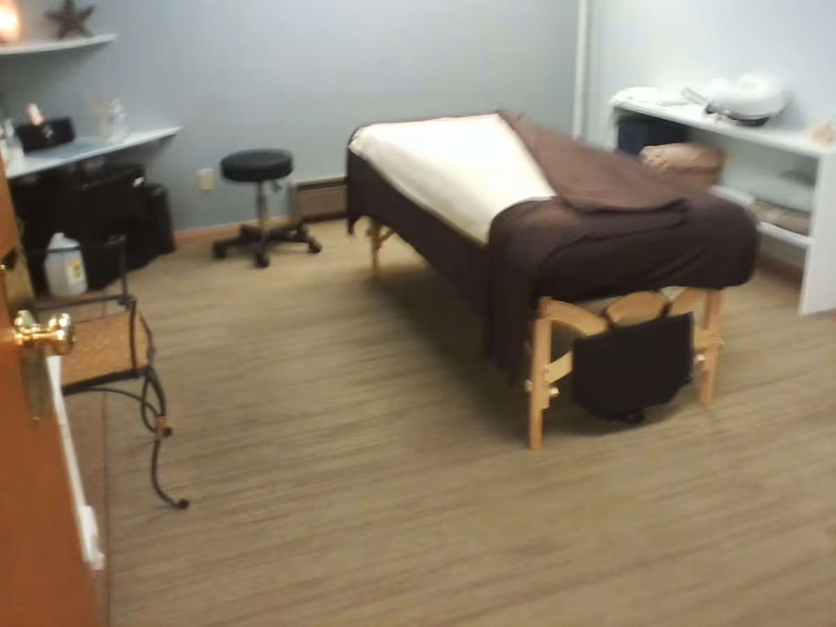 A small treatment room with a massage table, stool, and shelves of supplies.
