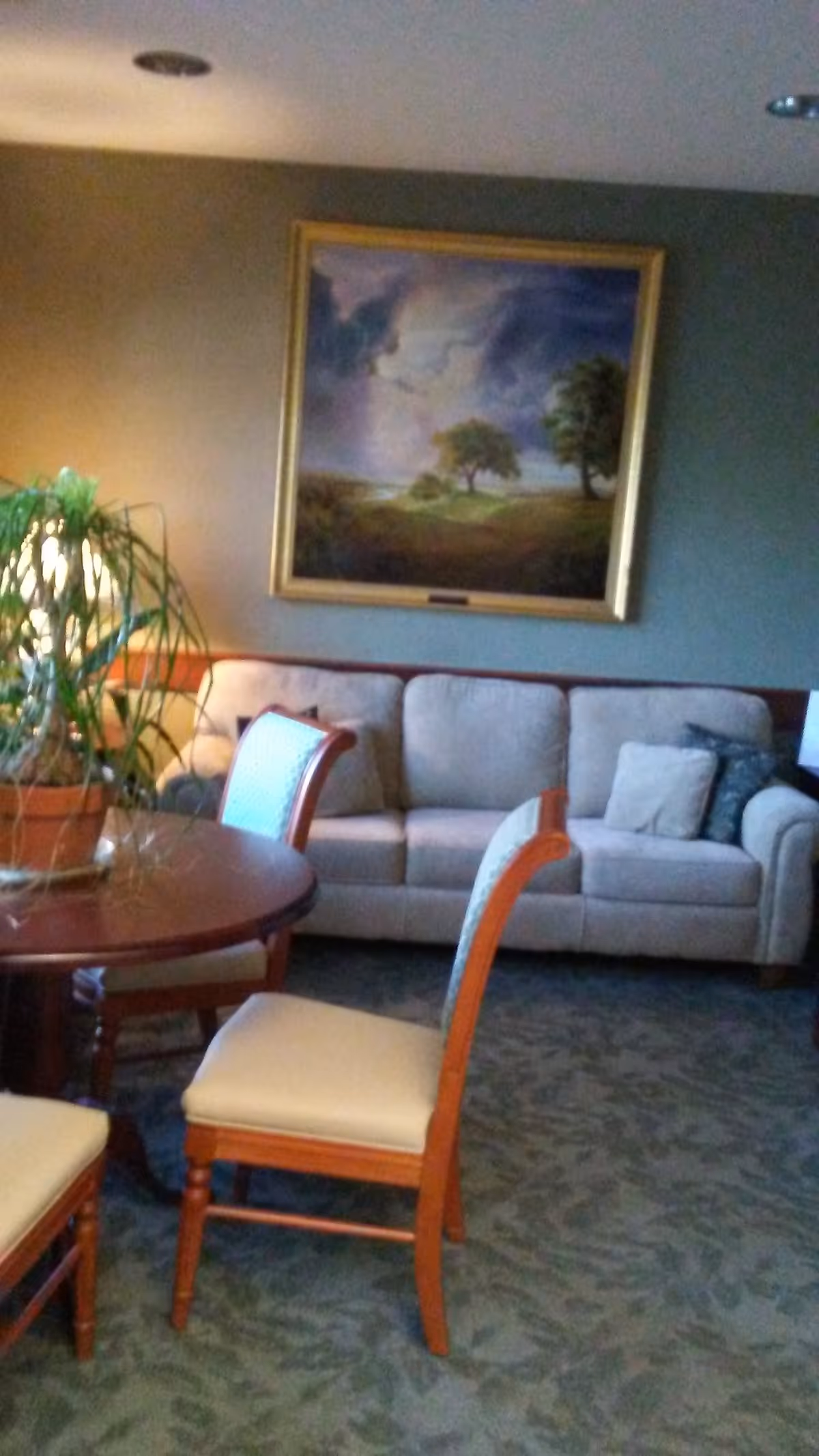 Cozy living room with beige sofa, round wooden table, chairs, plant, and framed landscape painting.