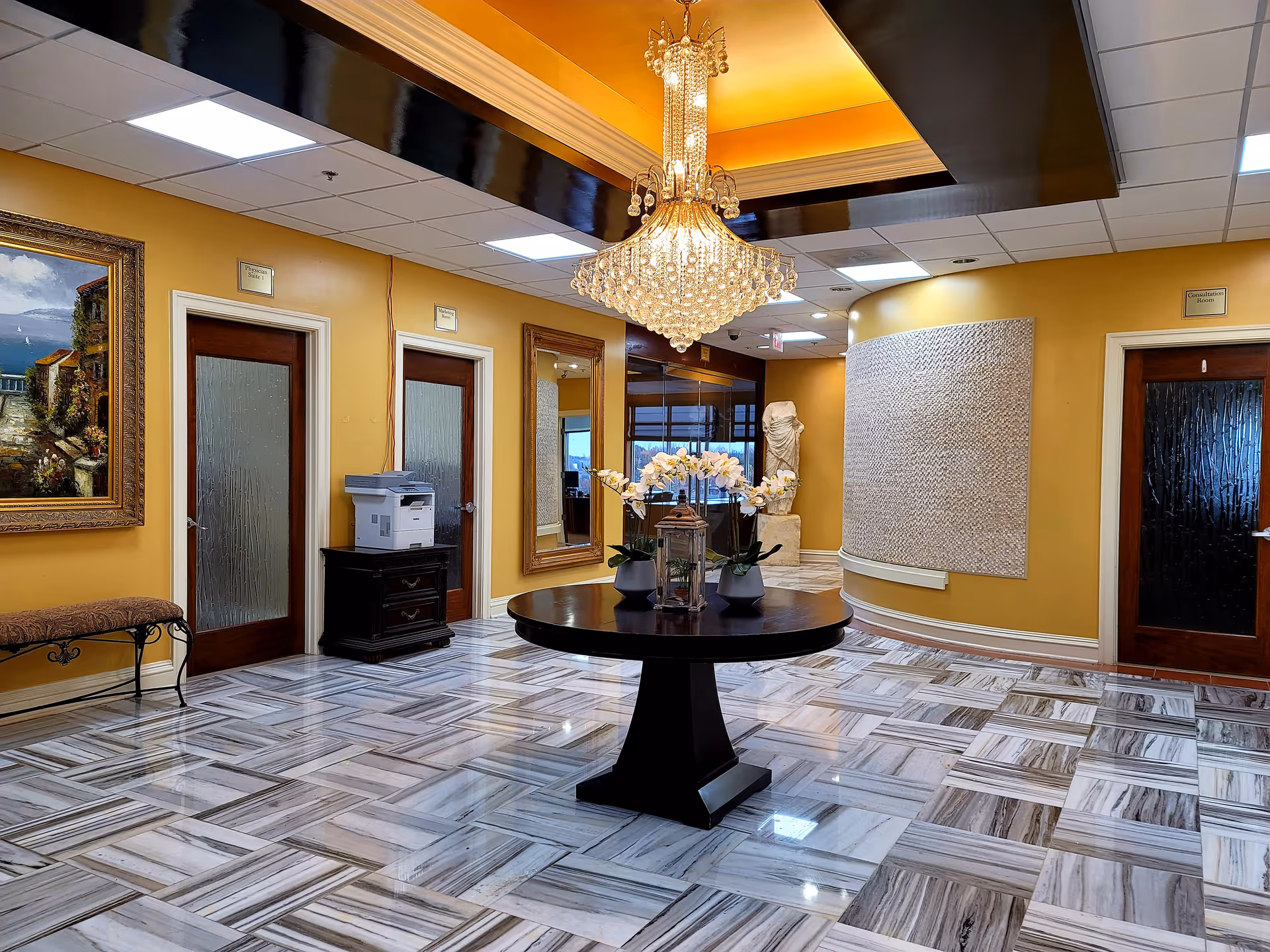 Luxurious yellow lobby with chandelier, round table, mirrors, marble floor, and potted orchids.