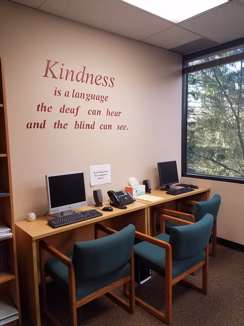 Office workspace with computers, phones, and teal chairs under a wall quote about kindness.