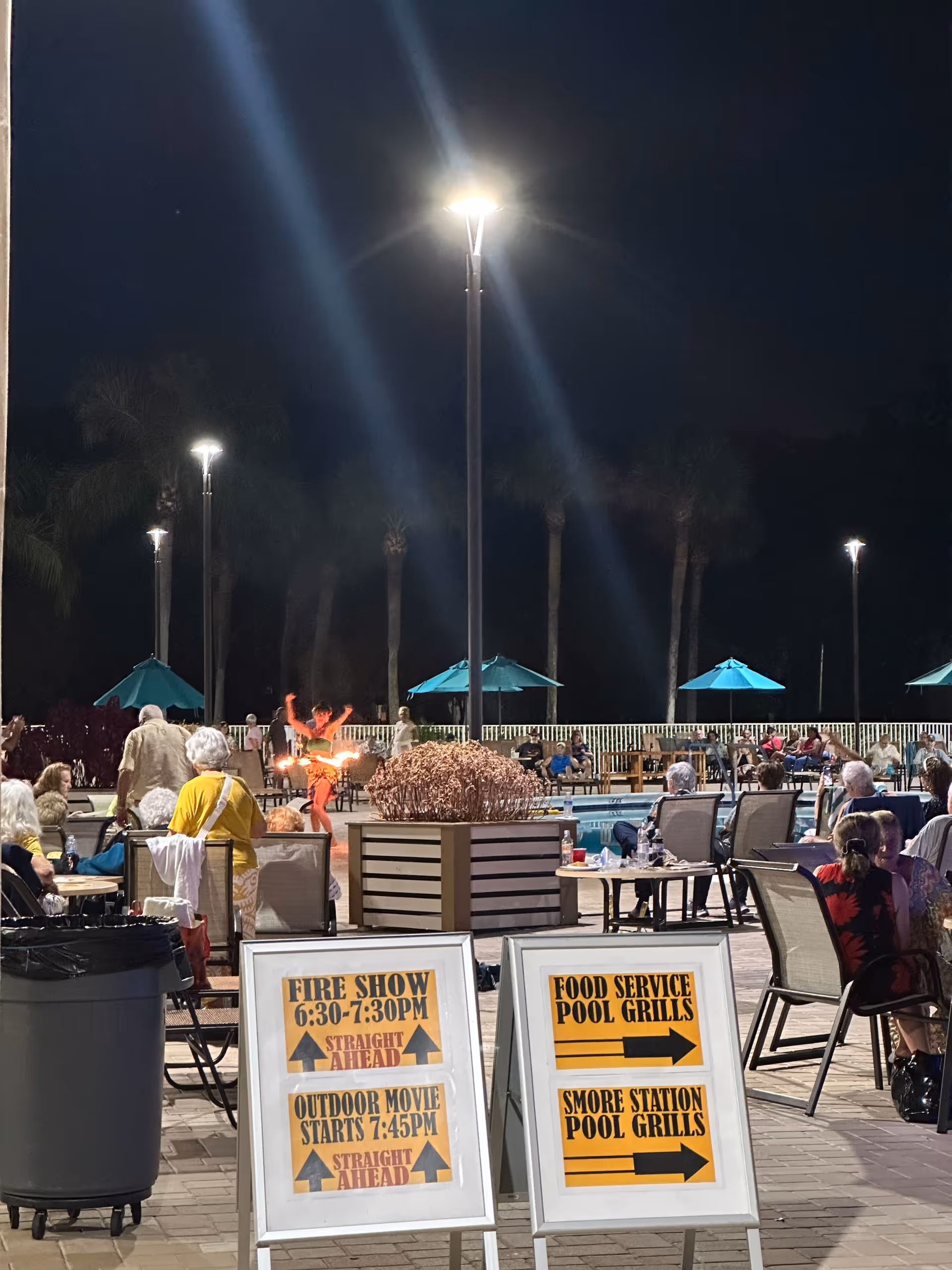 Nighttime outdoor pool area with diners and activity signs.