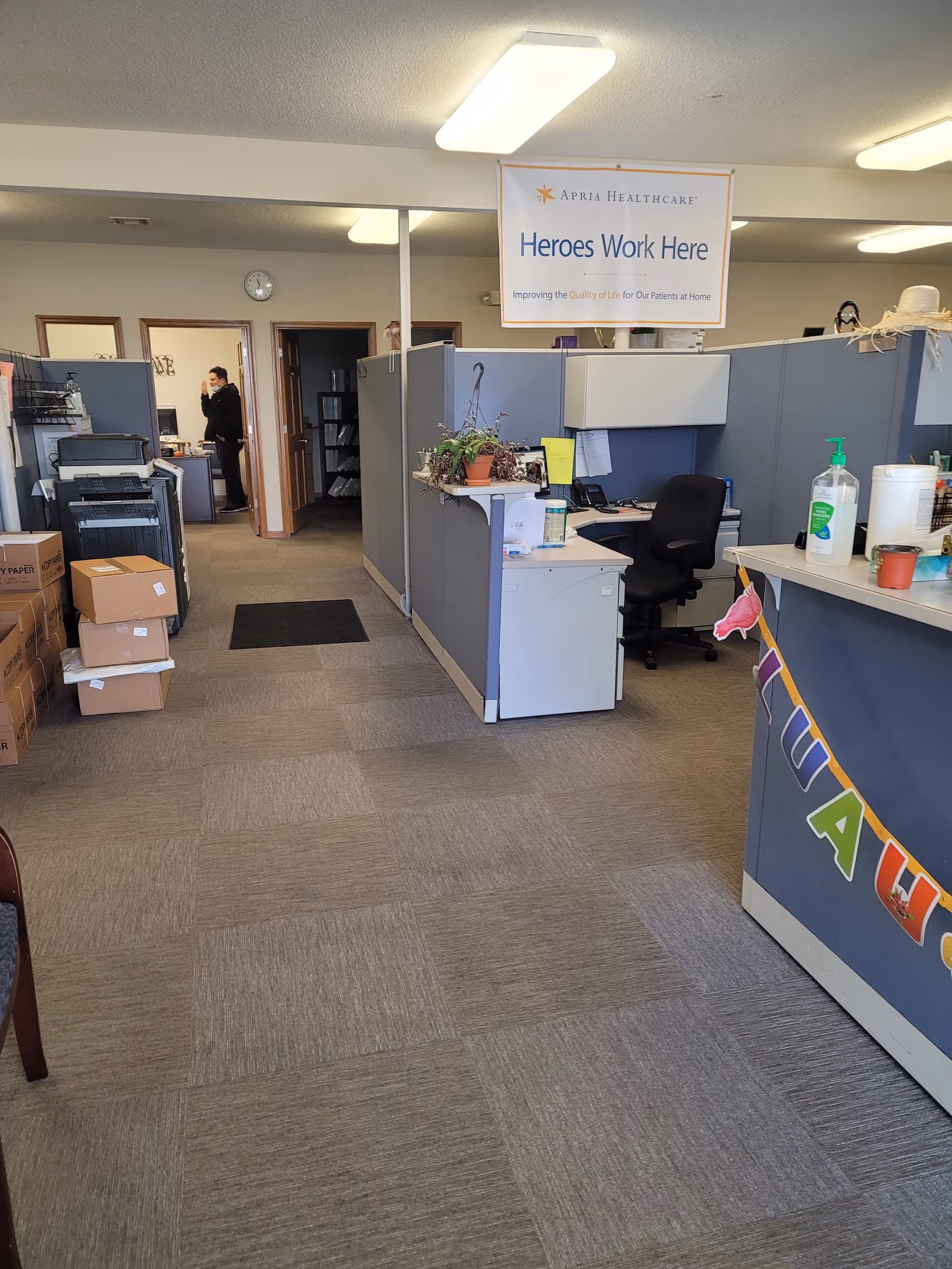 Apria Healthcare office with cubicles, desks, banner, plant decor, and a person in the background.