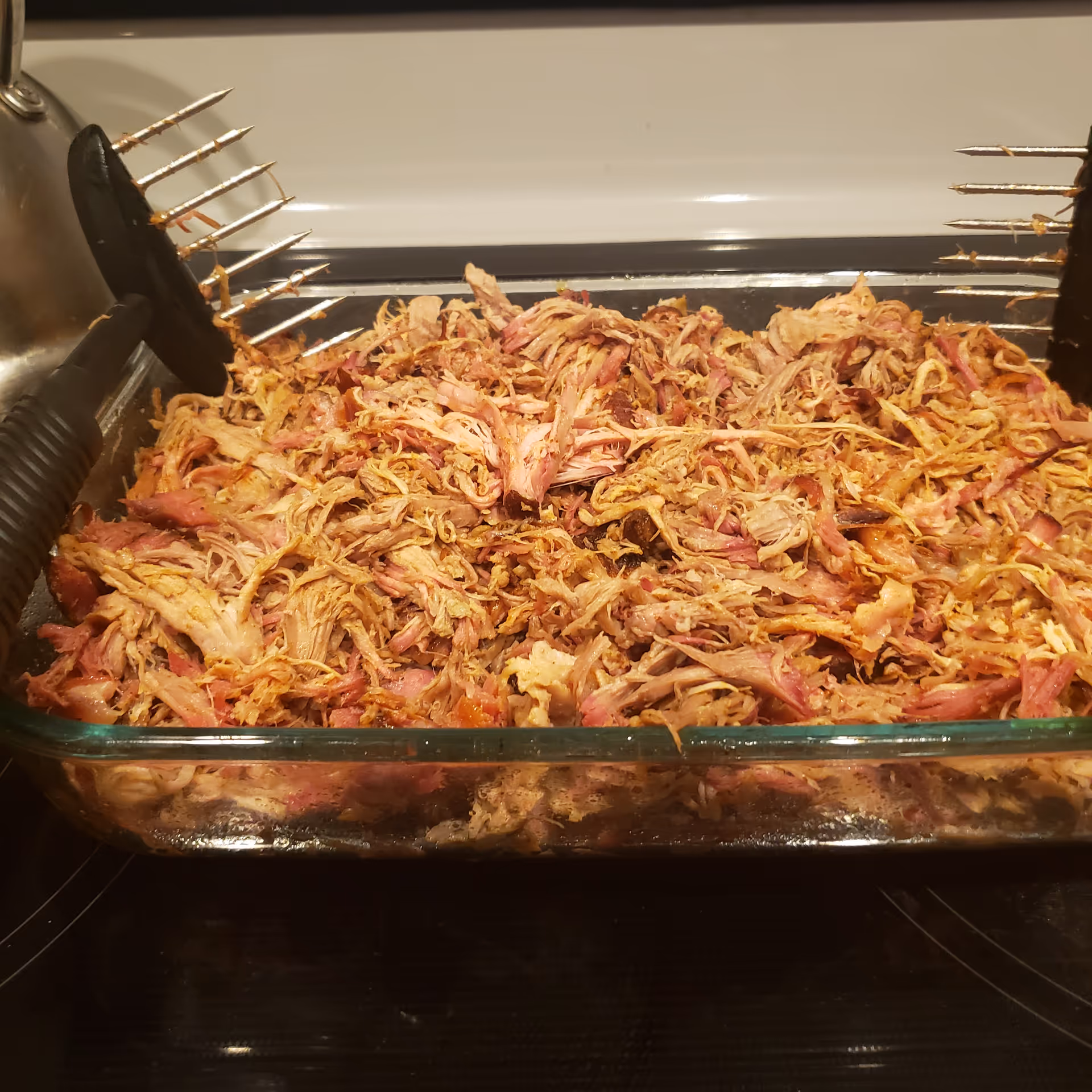 Glass baking dish of shredded pulled pork rests on a stove with tongs nearby.