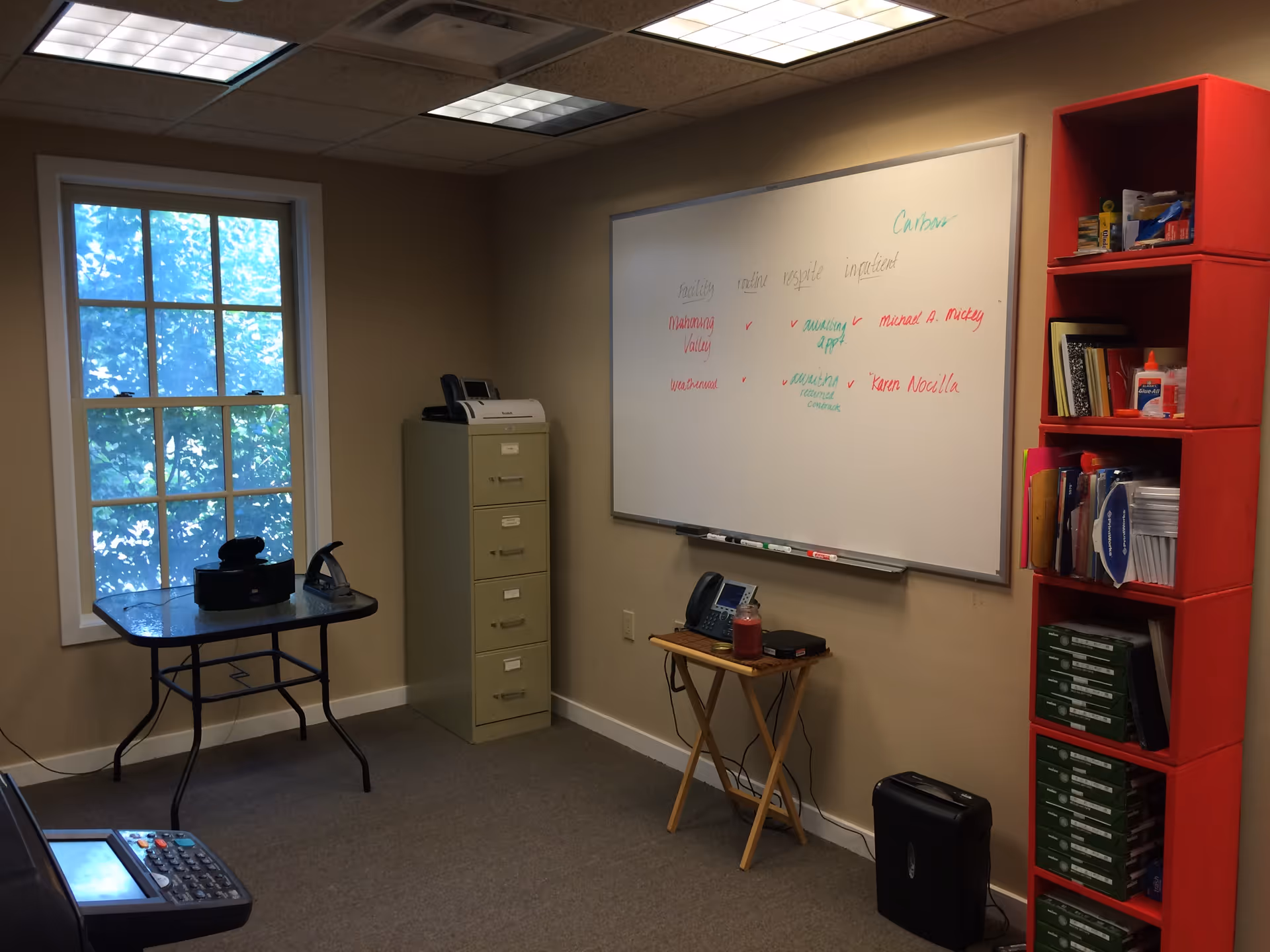 Interior office space with a whiteboard, red shelving, a filing cabinet, and a small table.