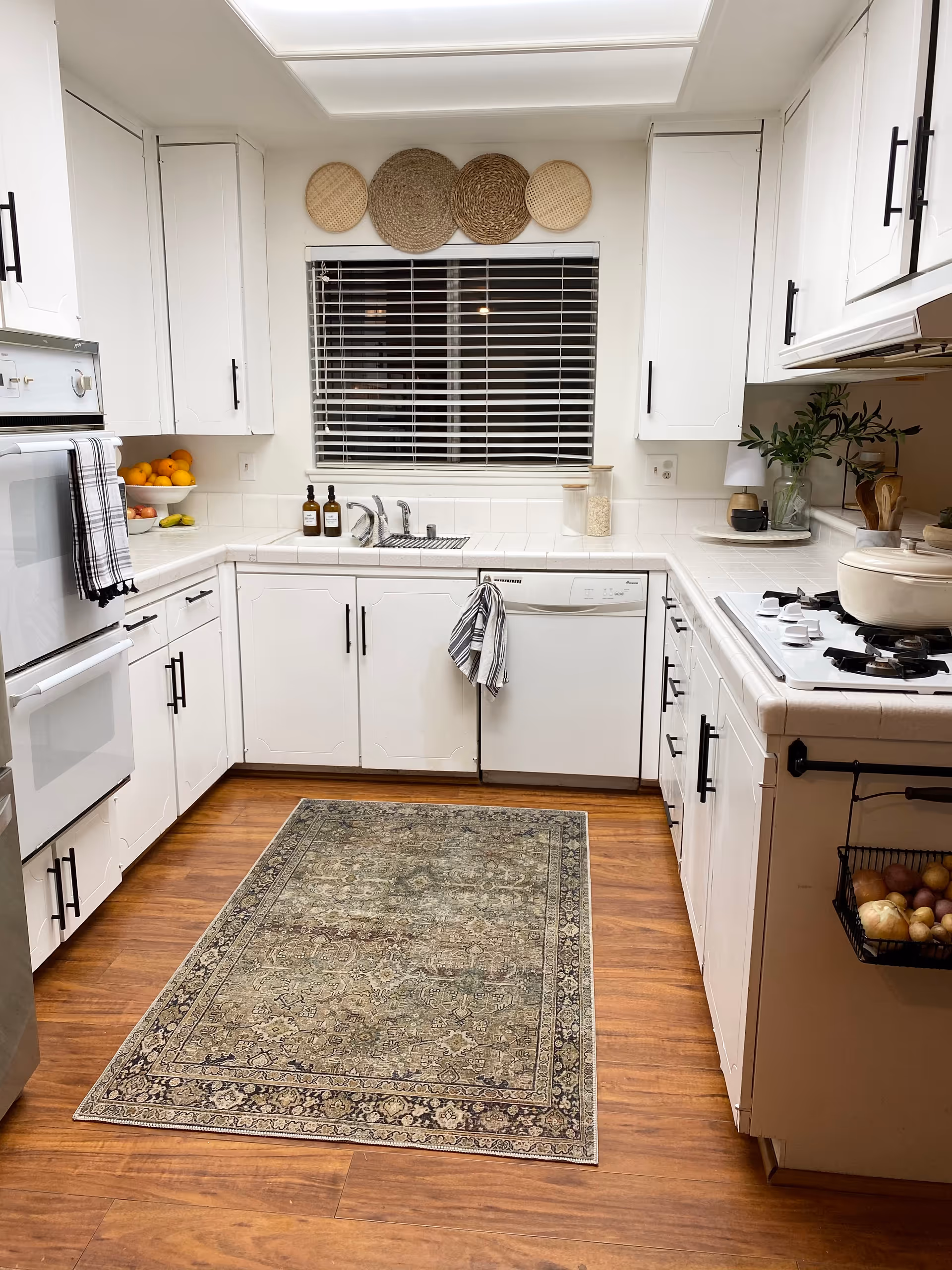 White kitchen with cabinets, sink, stove, window, and a patterned rug on wood floor.