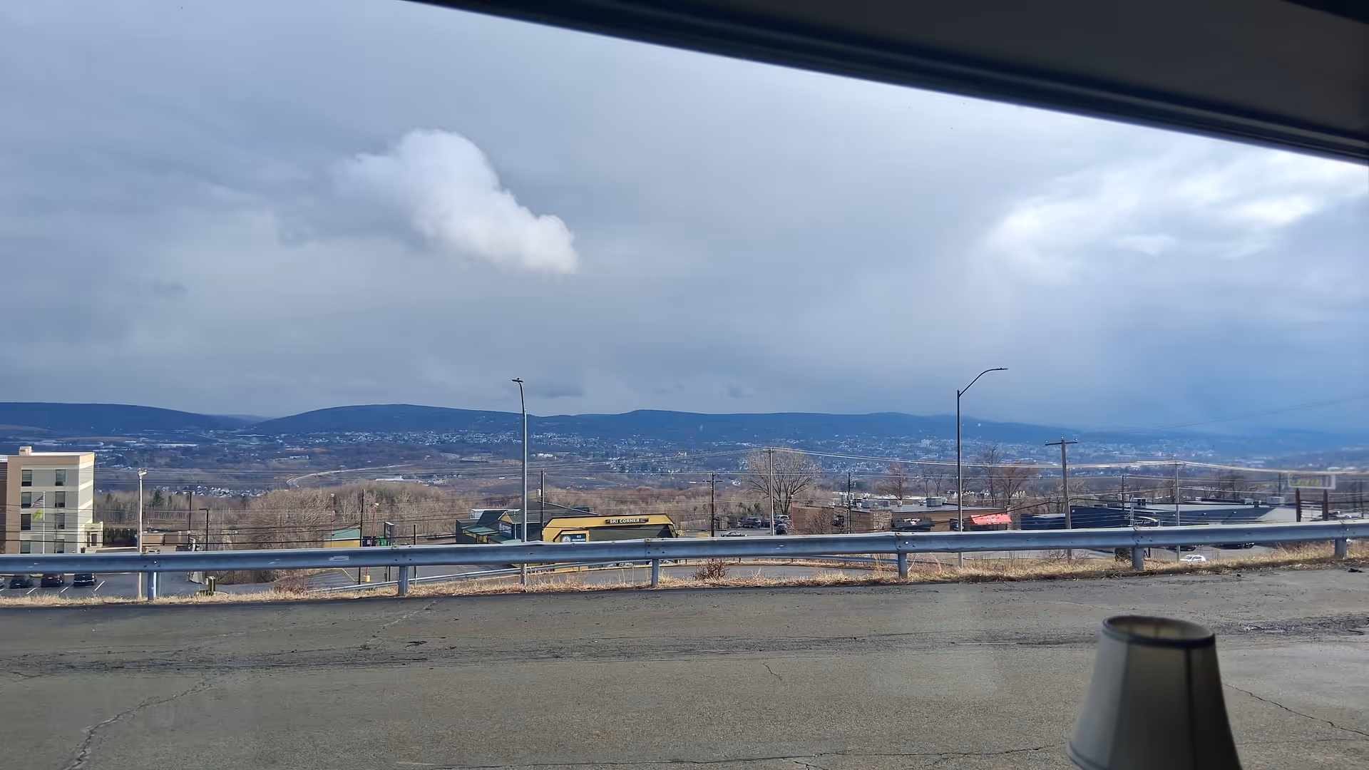View from a window shows a highway, guardrail, and distant hills under cloudy sky.