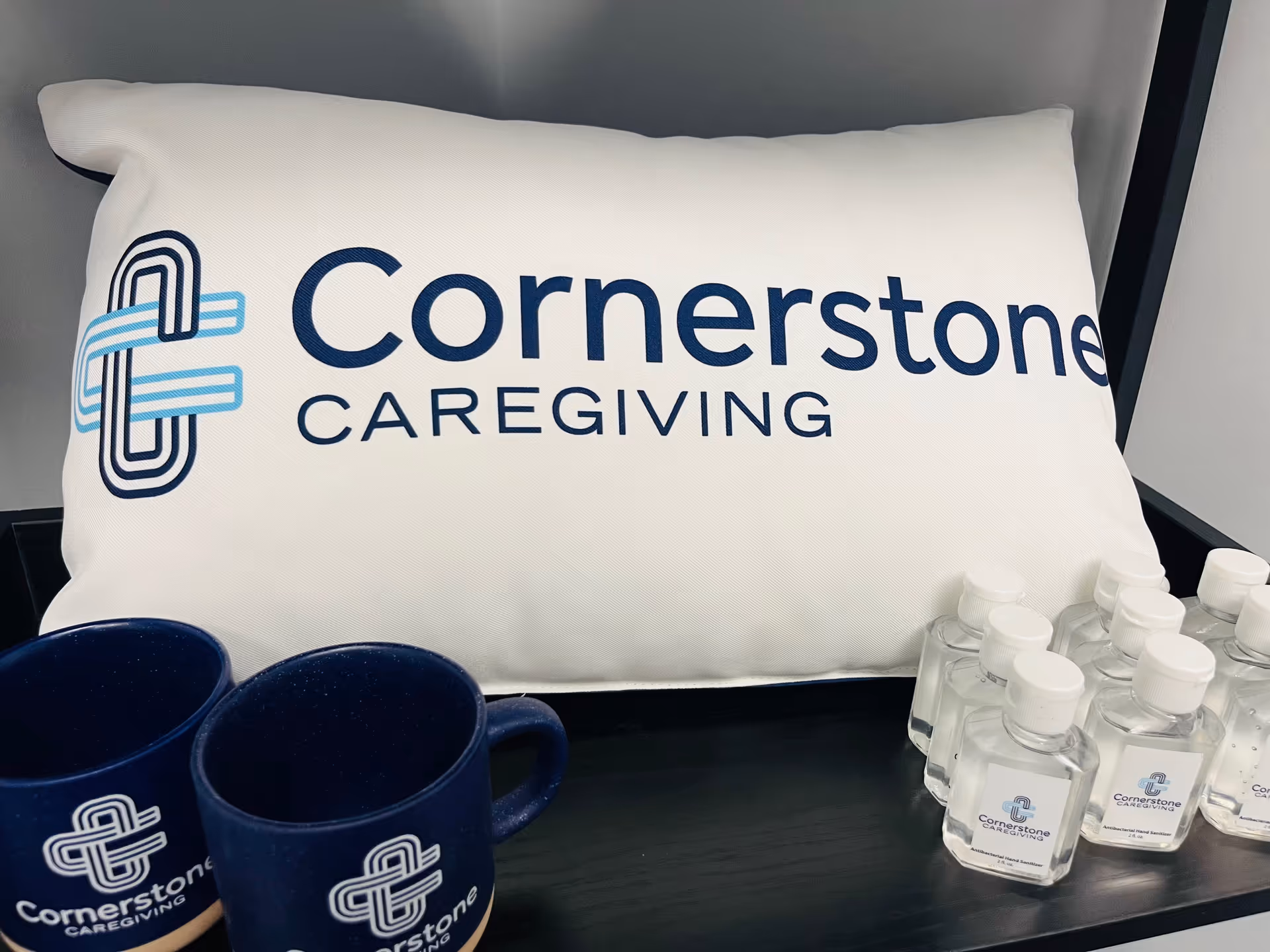 Pillow with Cornerstone Caregiving logo, blue mugs, and hand sanitizer bottles on a table.
