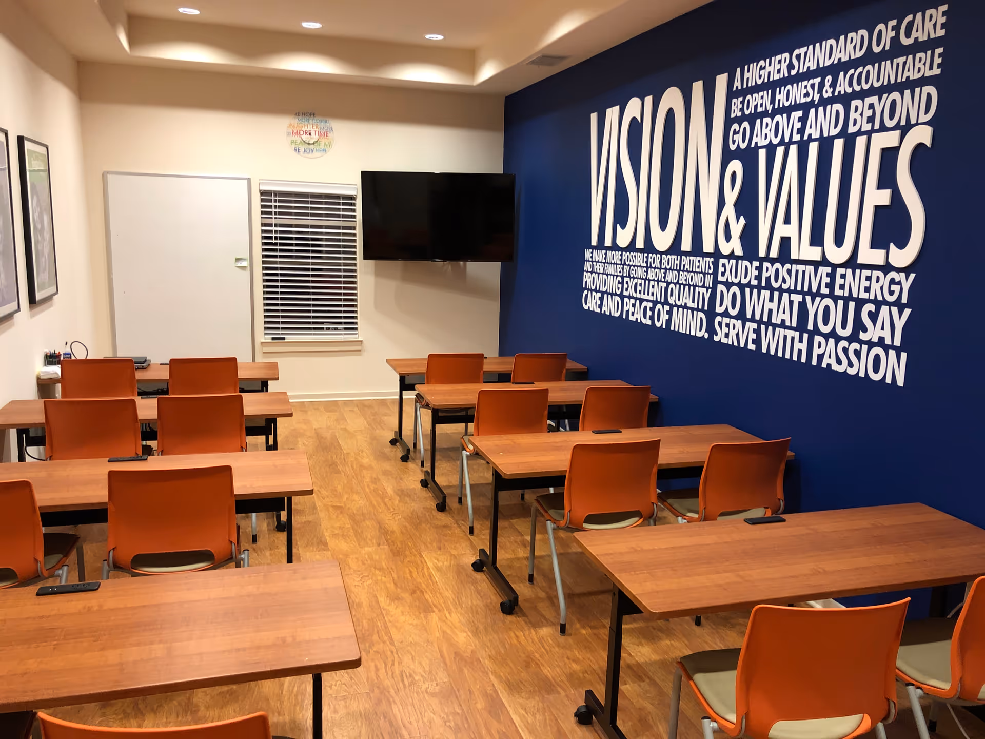 Bright classroom-style room with orange chairs, wooden tables, a TV, and a Vision & Values wall