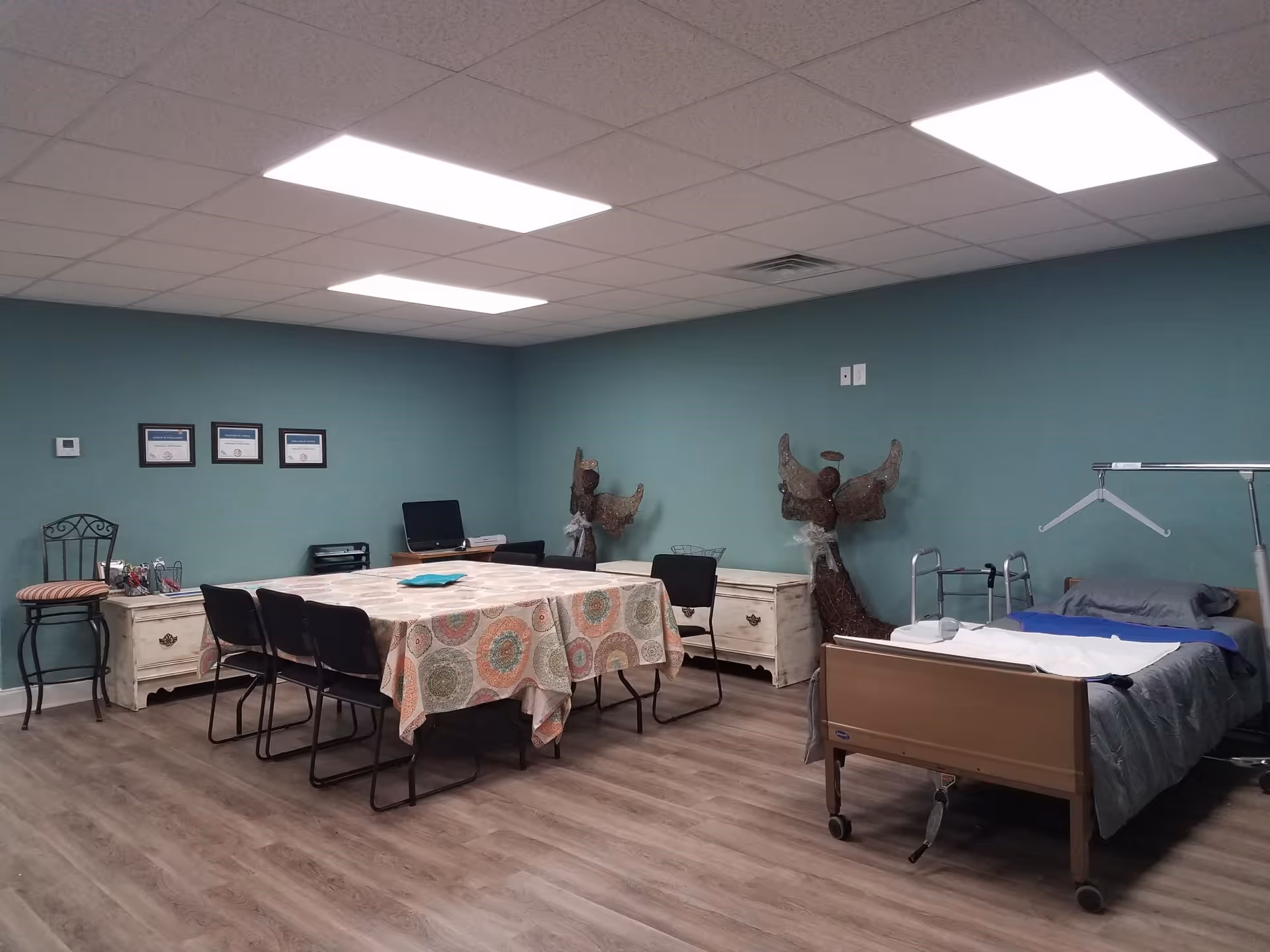 Teal-walled care facility room with a long dining table, chairs, and a hospital bed.