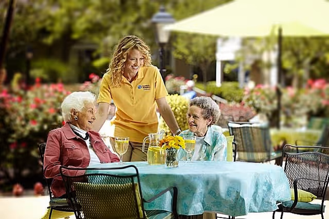 Caregiver chats with two elderly residents at an outdoor patio table.