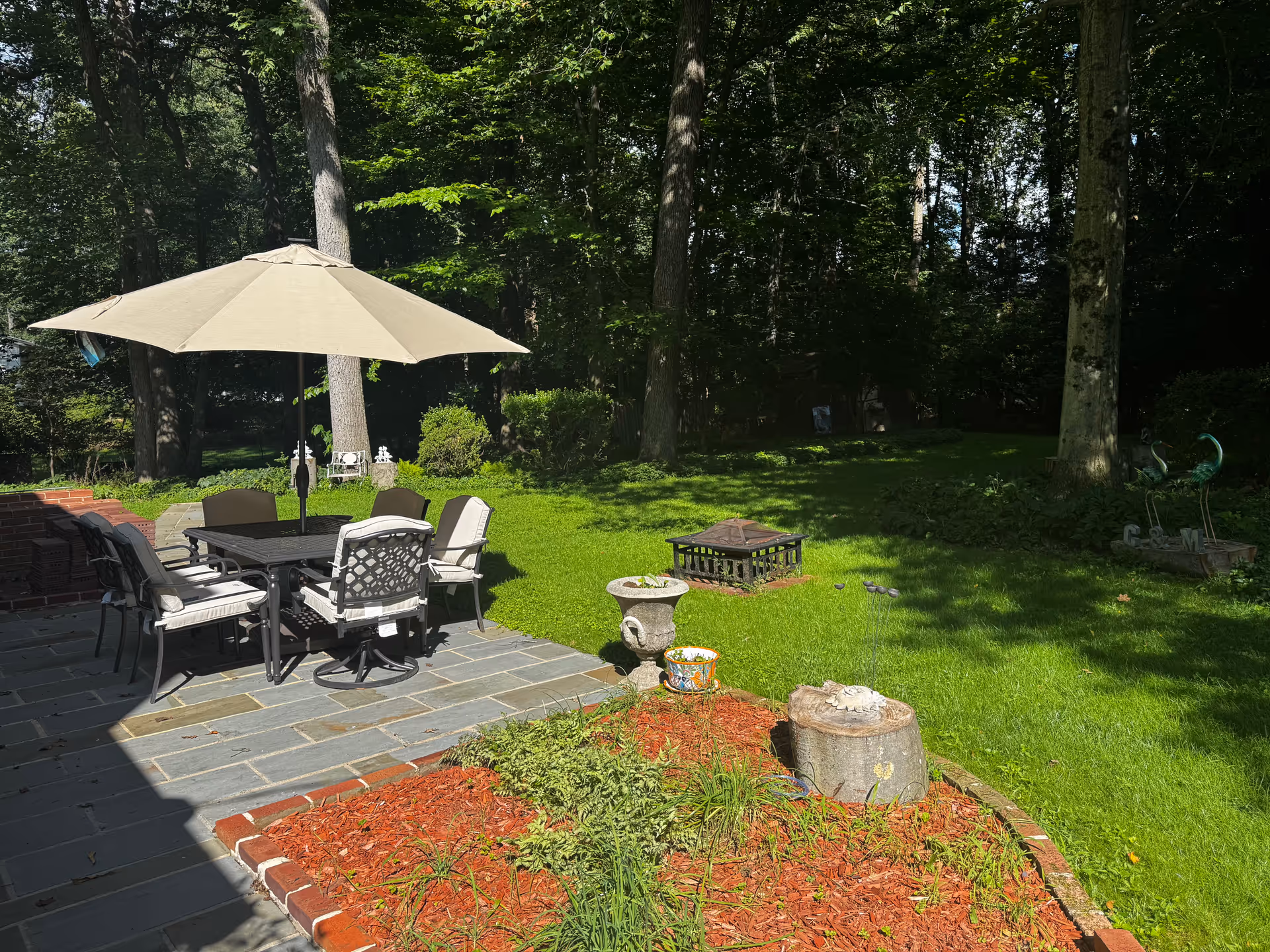 Outdoor patio area with table, chairs, umbrella, and surrounding green lawn and trees.