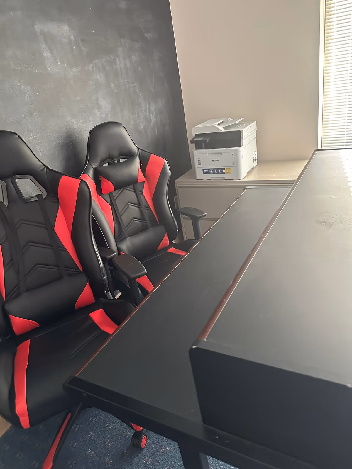 Office area with black and red gaming chairs beside a large desk and printer.