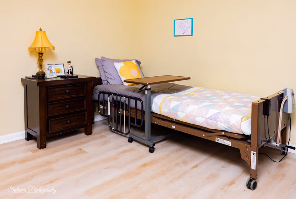 Bedroom with adjustable hospital bed, dresser, and pastel quilt in yellow-walled room.