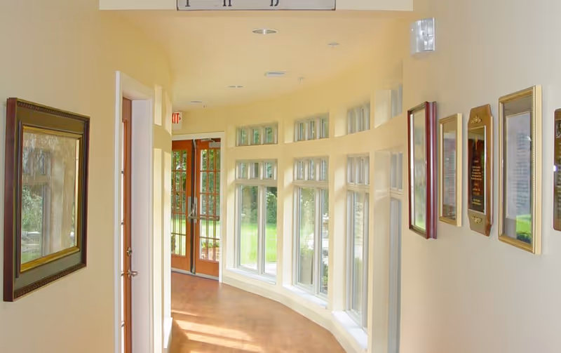 Bright curved hallway with large windows, wall plaques, and a door at the end.