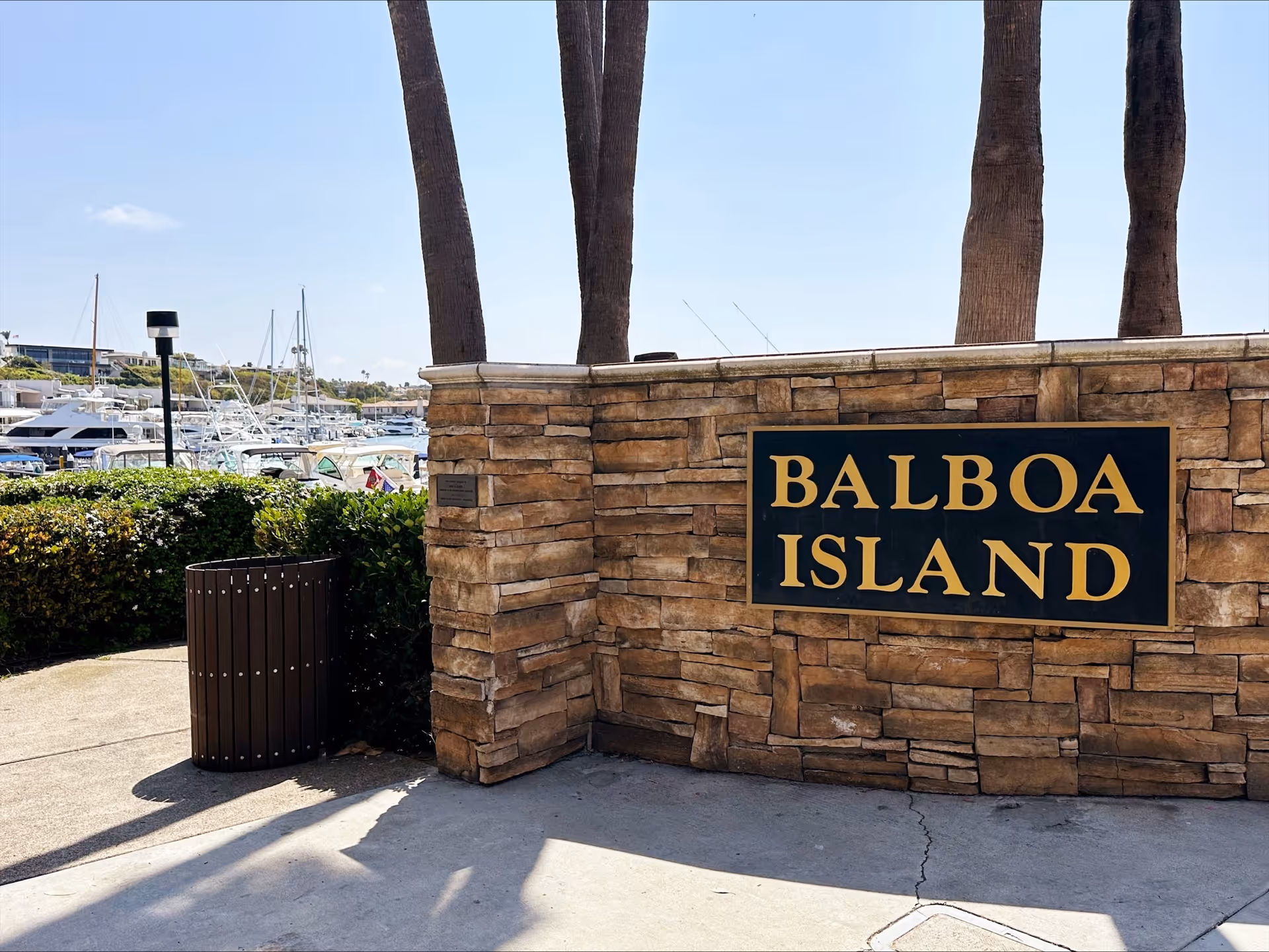 Stone wall with a Balboa Island sign, palm trees, and a marina with boats.