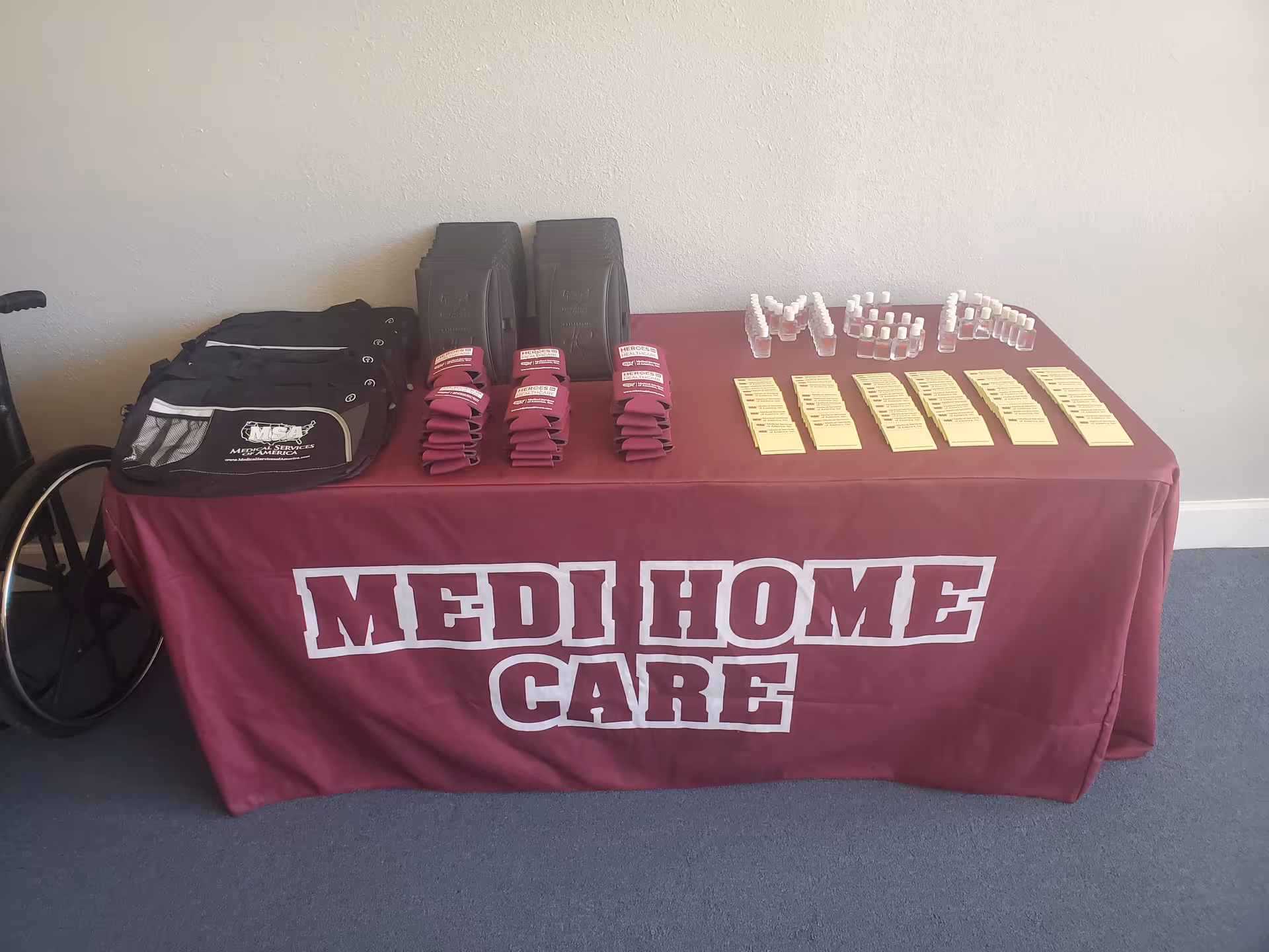 Indoor table with Medi Home Care branded burgundy cloth, backpacks, and promotional items.
