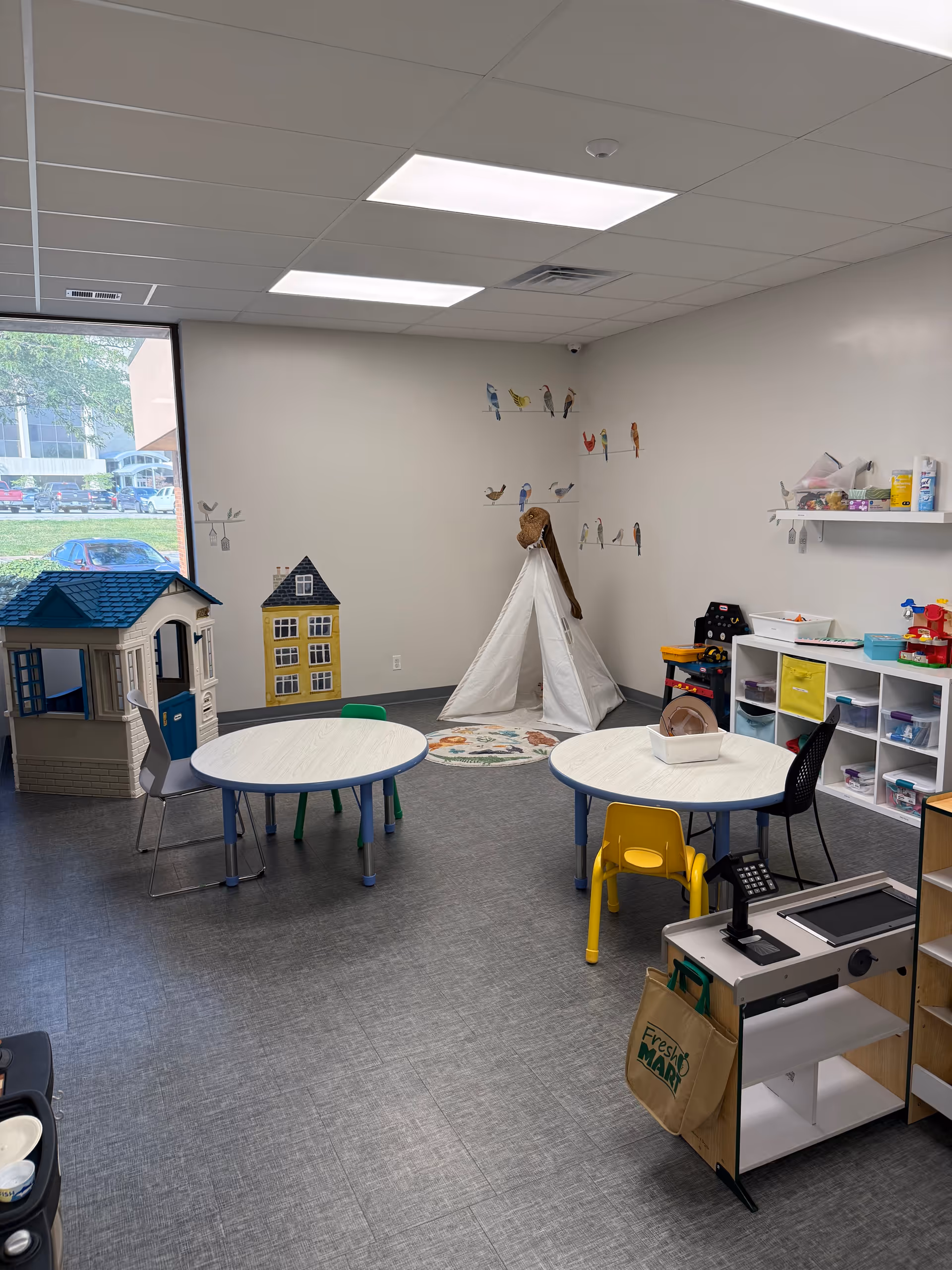 Interior children's activity room with two toy houses, a teepee, tables, and storage shelves.