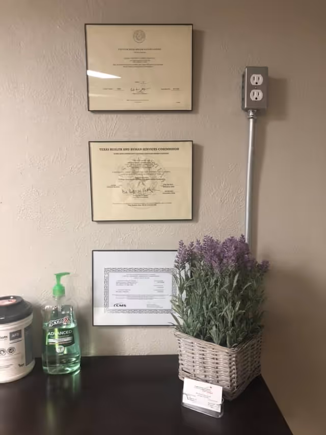 Interior wall with three certificates and a lavender plant on a dark surface next to sanitizer.