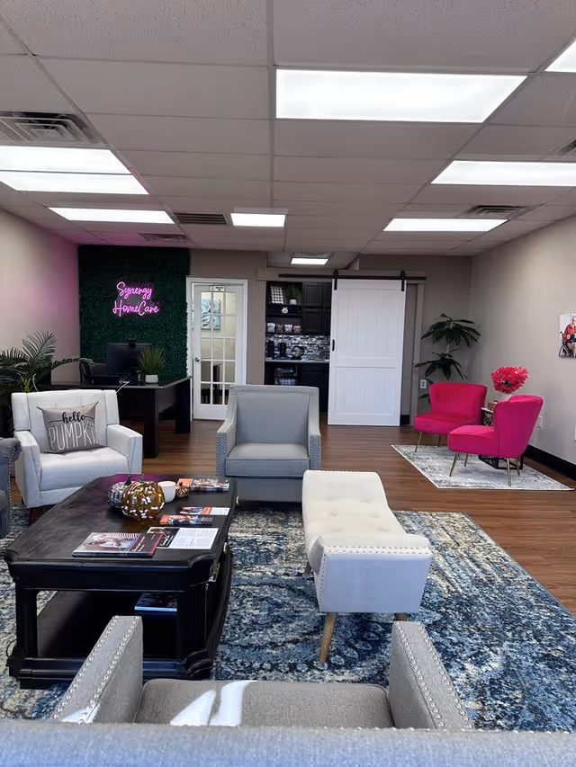 Modern lobby lounge with grey chairs, a coffee table, rug, plants, and a neon Synergy sign.