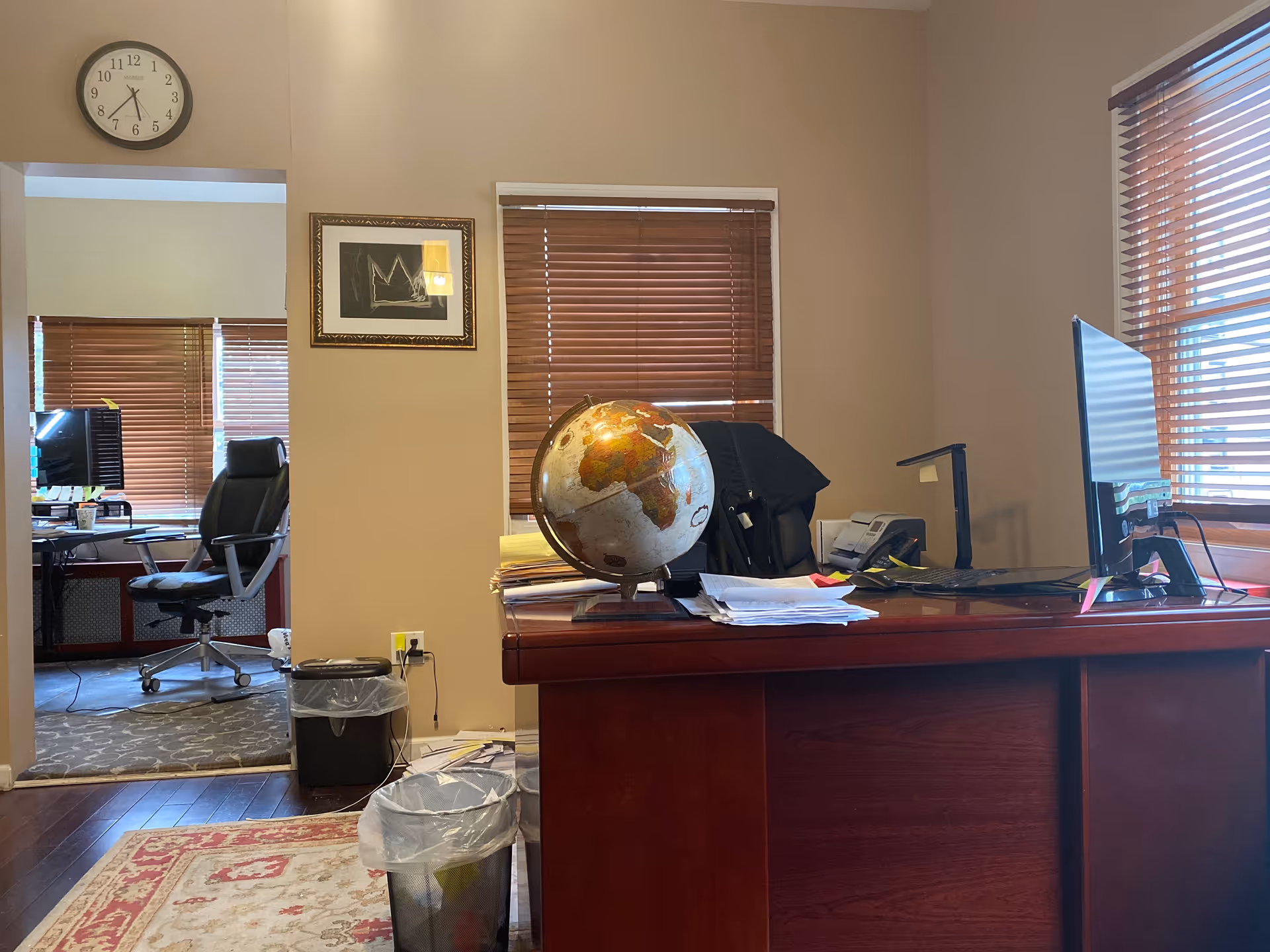 Office reception area featuring a wooden desk, globe, computer, blinds, clock, and doorway.