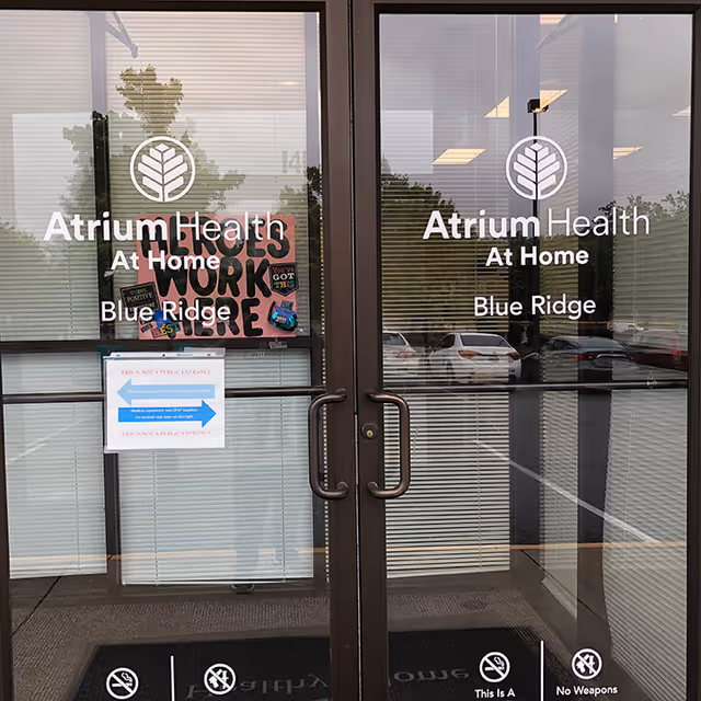 Glass entrance doors display Atrium Health At Home Blue Ridge with reflections of parked cars