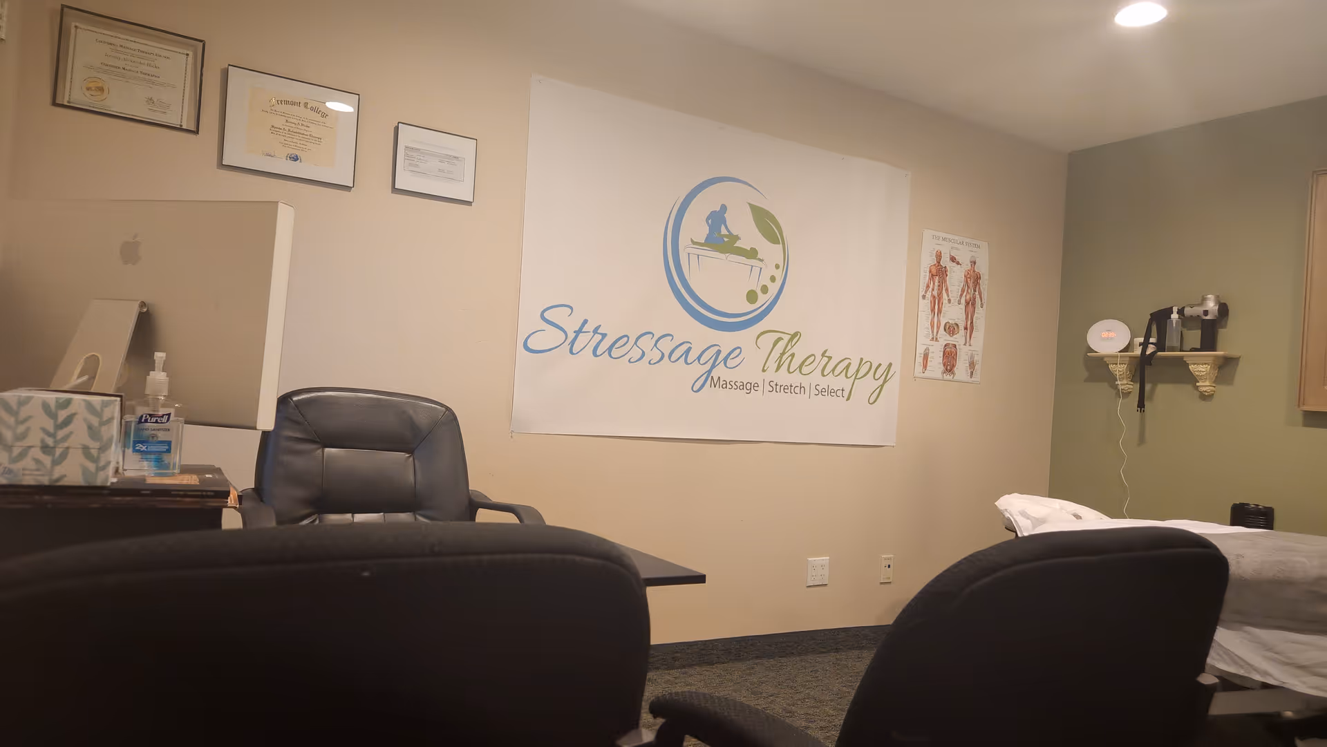 Clinic room featuring Stressage Therapy banner, chairs, certificates, a treatment table, and hand sanitizer.