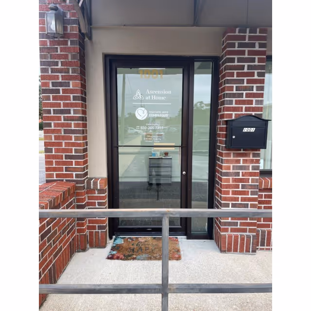 Brick exterior entrance with glass door reading Ascension at Home; brick walls and railing.