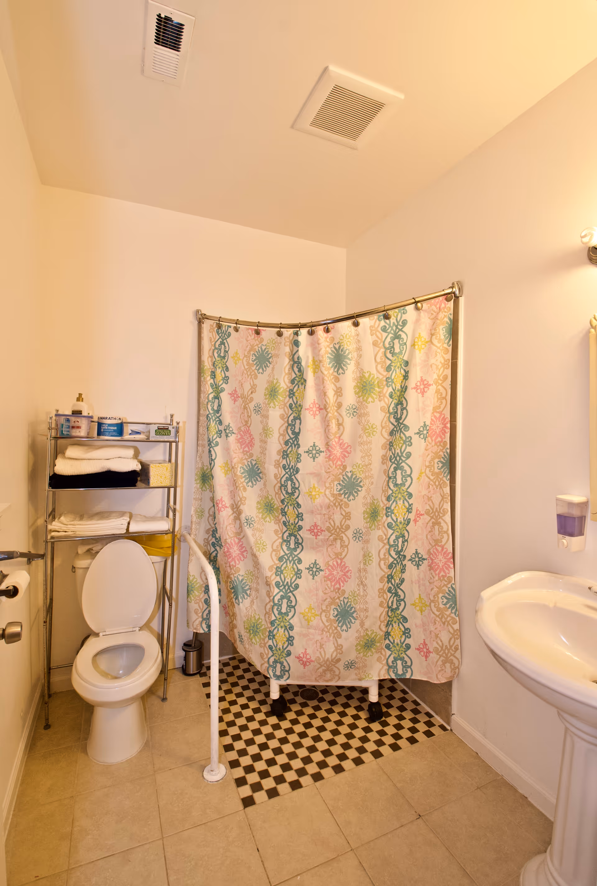 Accessible bathroom with toilet, colorful shower curtain, sink, and metal shelf holding towels.