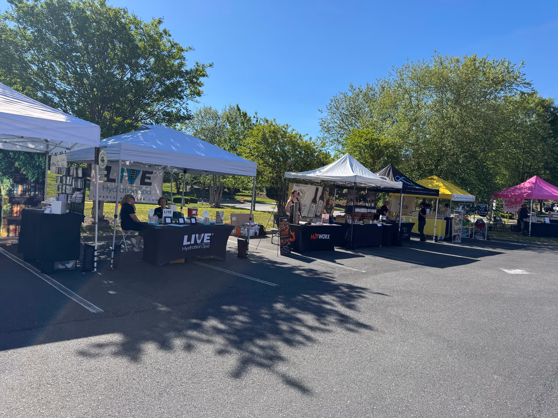 Row of vendor tents set up in a parking lot with tables and shade under trees.