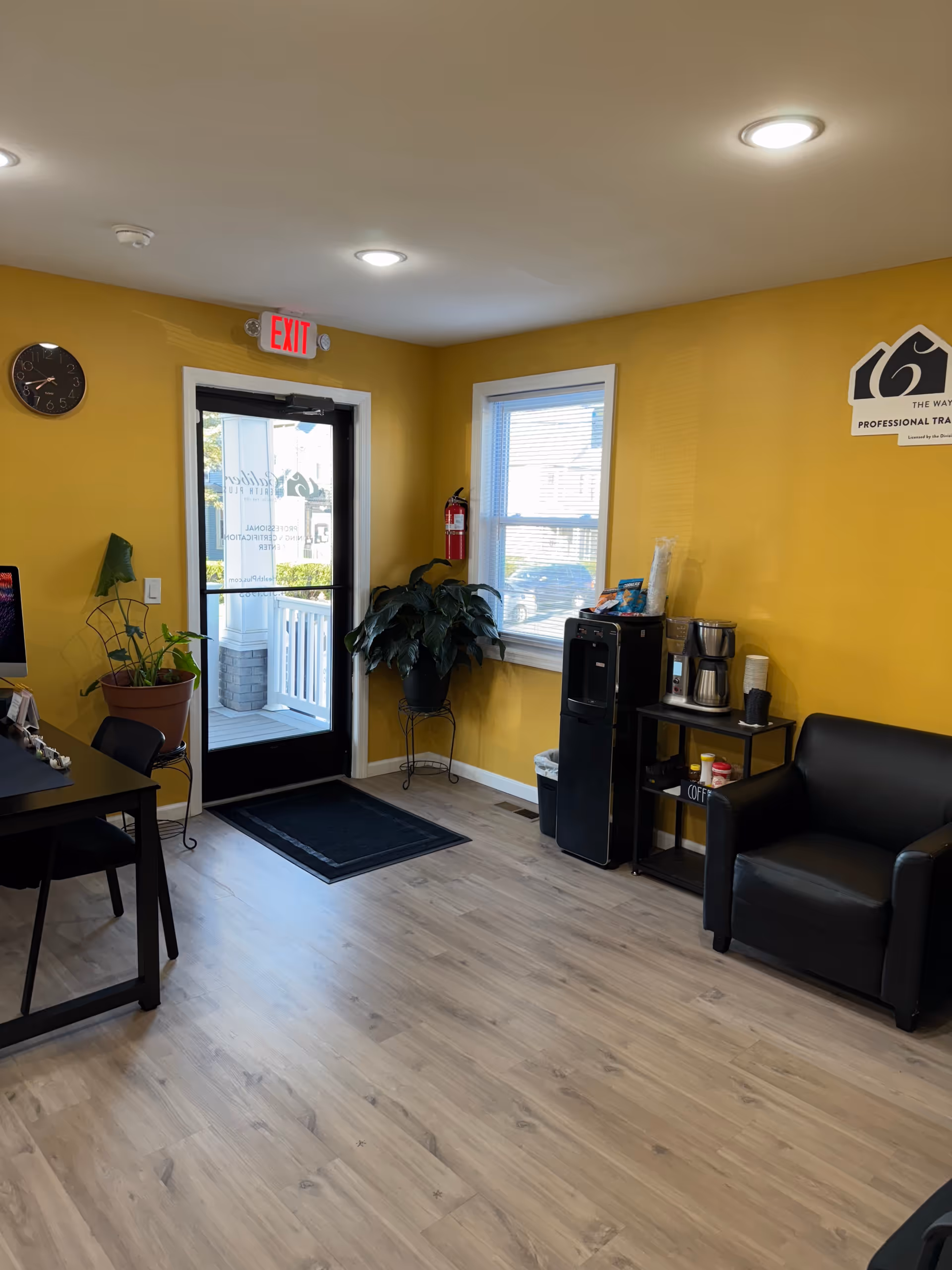 Bright yellow lobby area with exit door, window, plants, coffee station, and black seating.