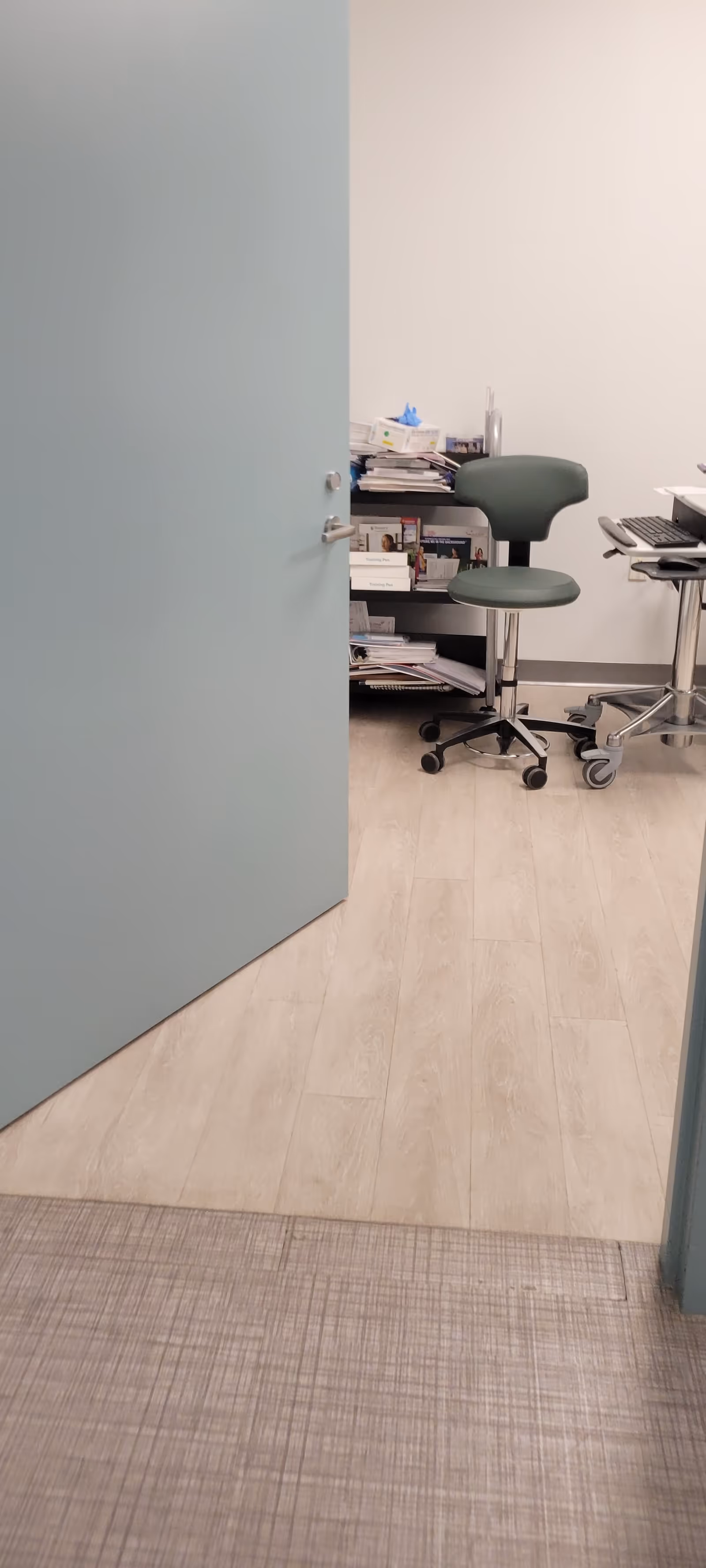Open teal door reveals a medical office area with a rolling chair and computer.