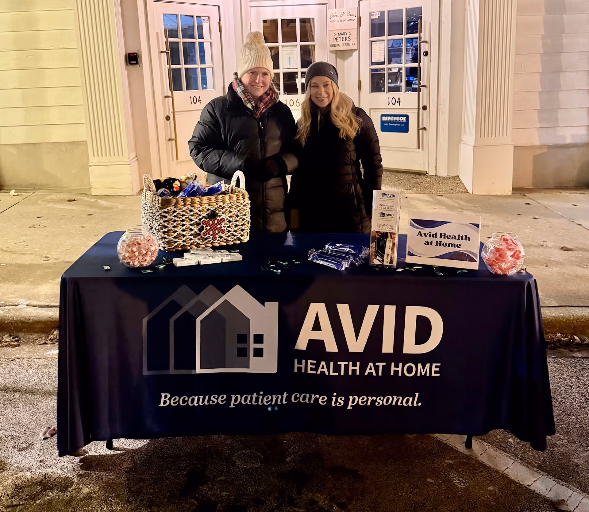 Two people stand behind an outdoor promotional table for AVID Health at Home.
