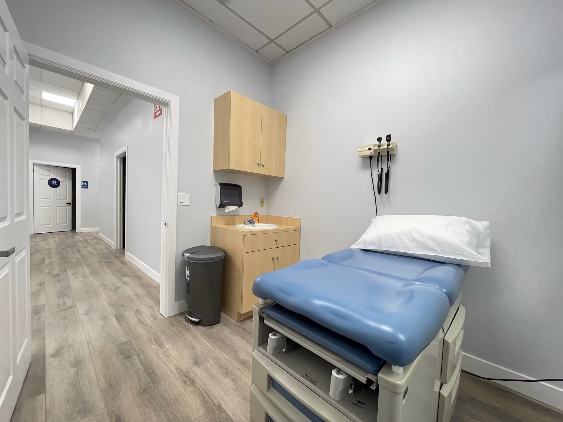 Clinic-style examination room with blue examination chair, sink, and wooden cabinets.