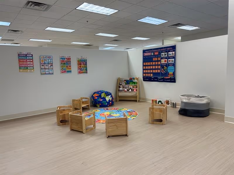 Interior therapy playroom with small wooden chairs, colorful rug, posters, and toy storage.