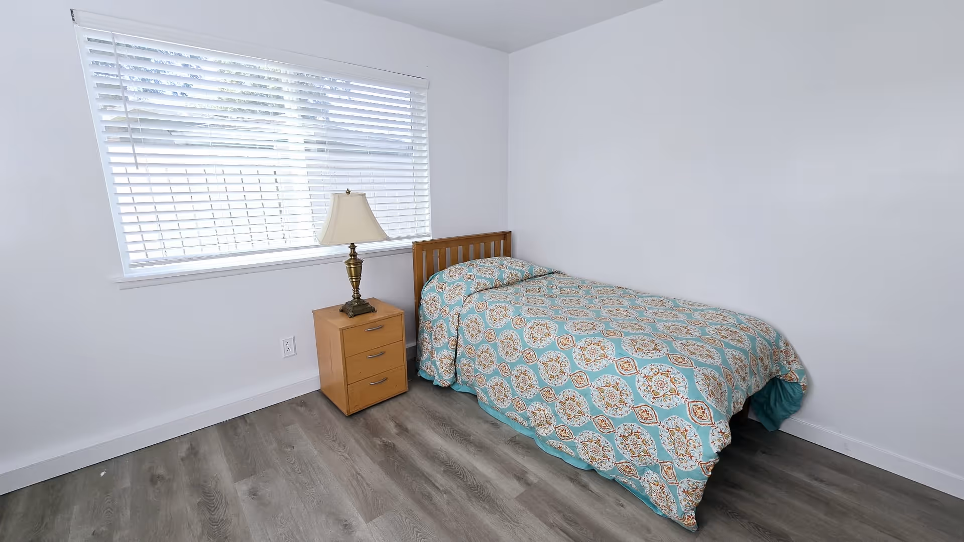Simple bedroom with single bed, nightstand, lamp, wood floor, and window blinds.