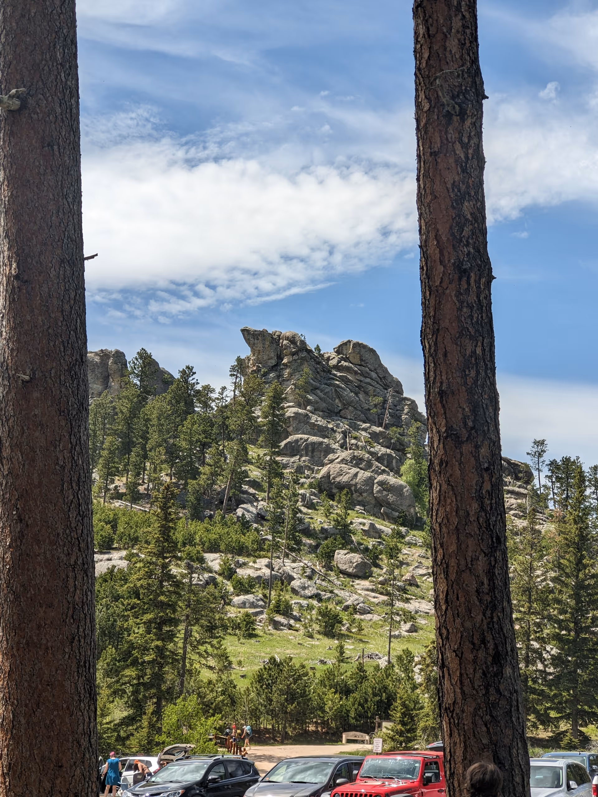 Tall trees frame a rocky hill with people and parked cars under a blue sky.
