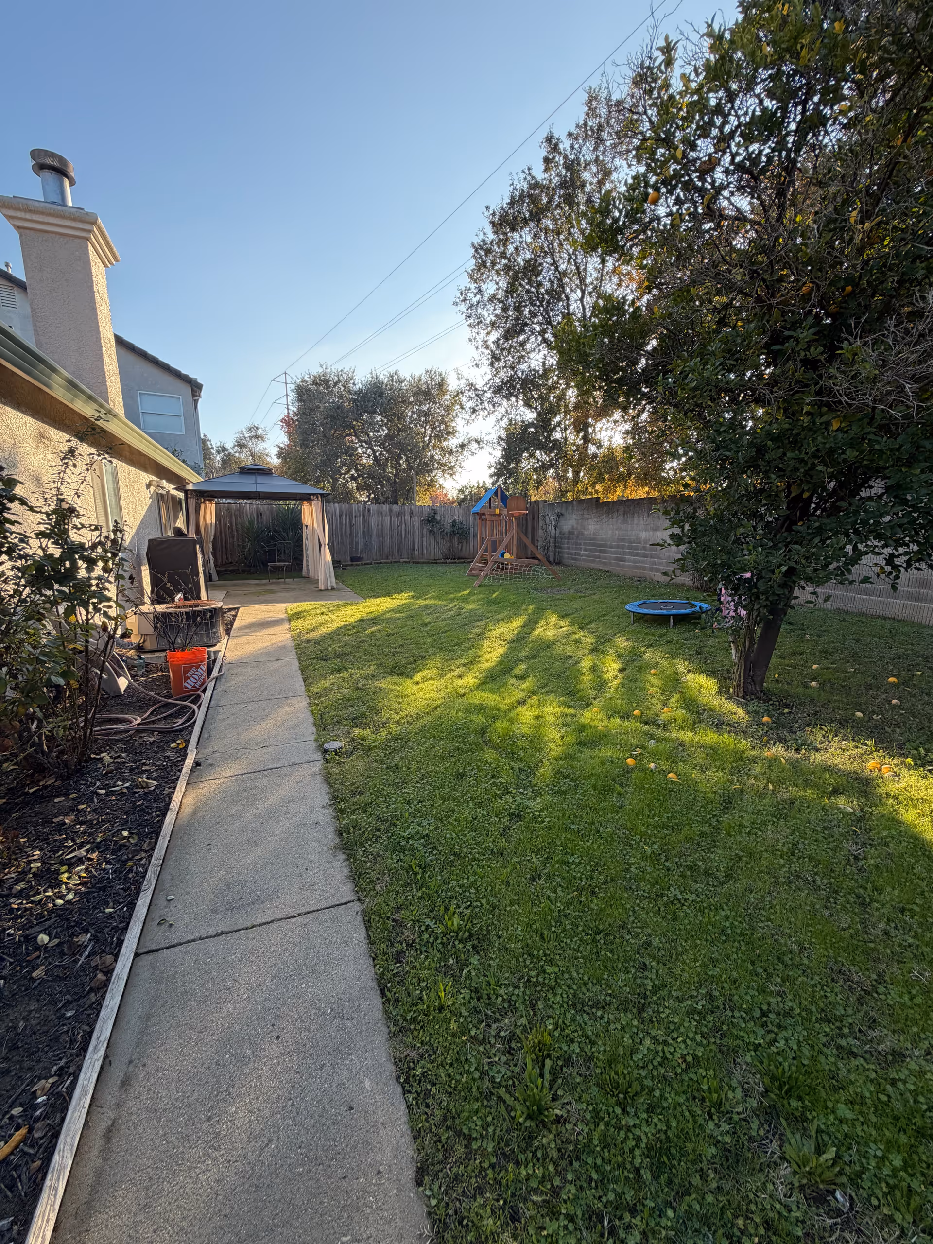 Sunny backyard with lawn, a concrete path, gazebo, playset, and a trampoline.