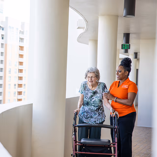 An elderly woman uses a walker while a caregiver assists along an outdoor corridor.