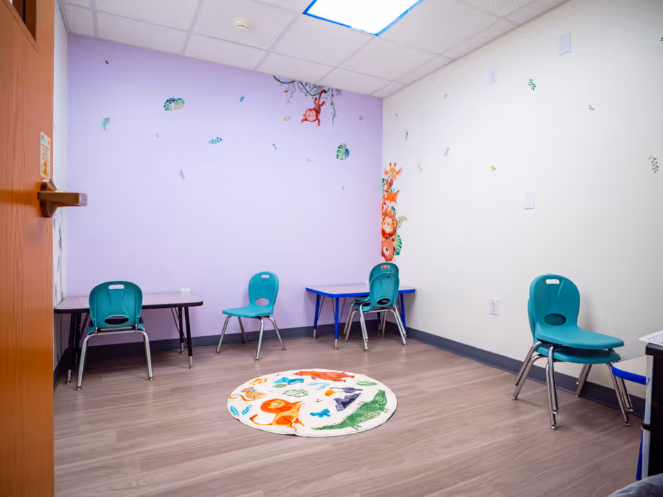 A child-friendly activity room with teal chairs, tables, purple wall, and a colorful circular rug.