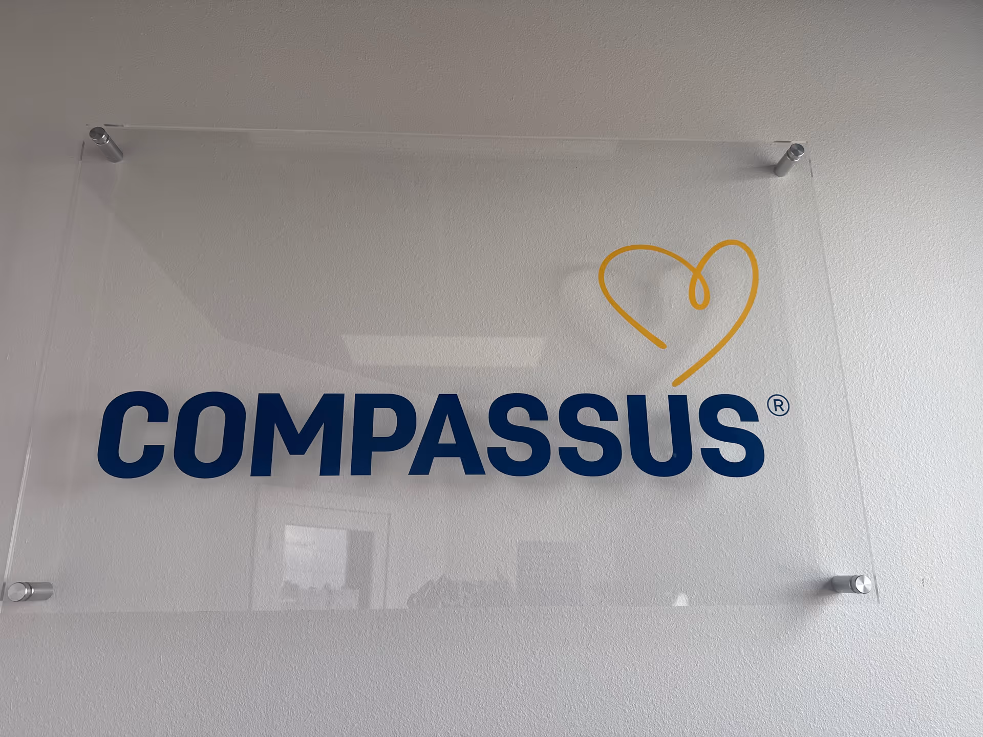 Clear acrylic sign reading COMPASSUS with yellow heart logo on white wall.