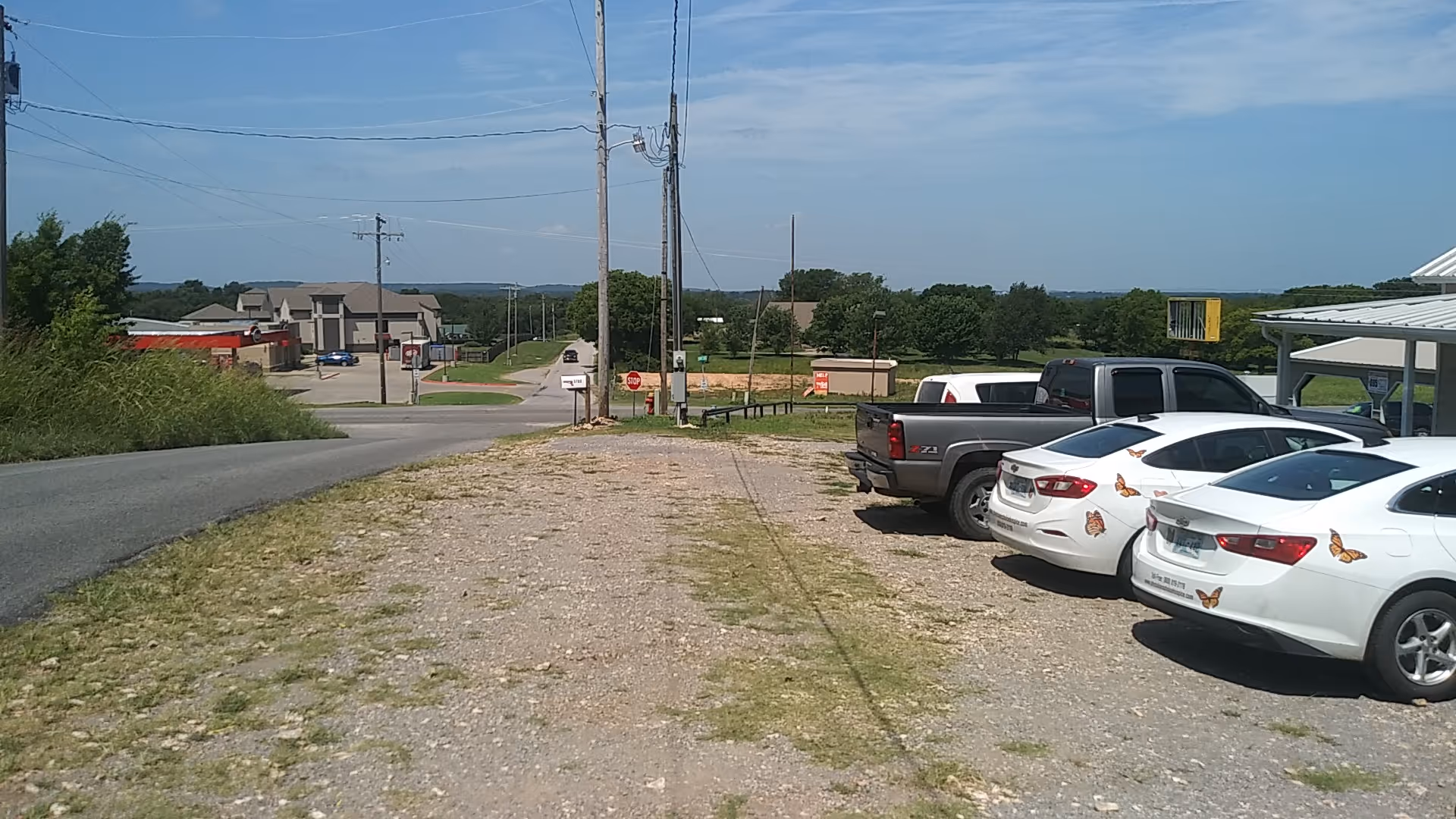 Exterior scene of a gravel parking area with parked cars and distant buildings.