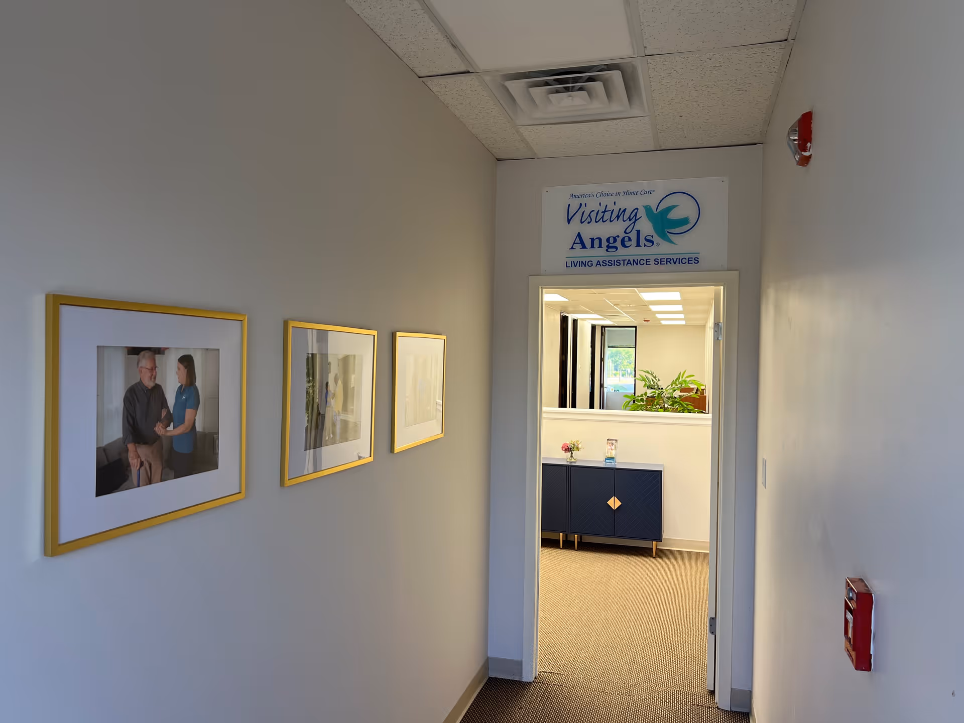 Hallway of a Visiting Angels facility with framed photos and reception area visible through the doorway.