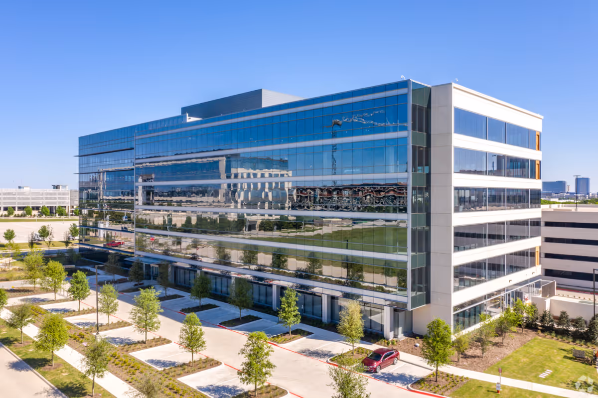 A modern glass-front office building with trees, parking, and clear blue sky.