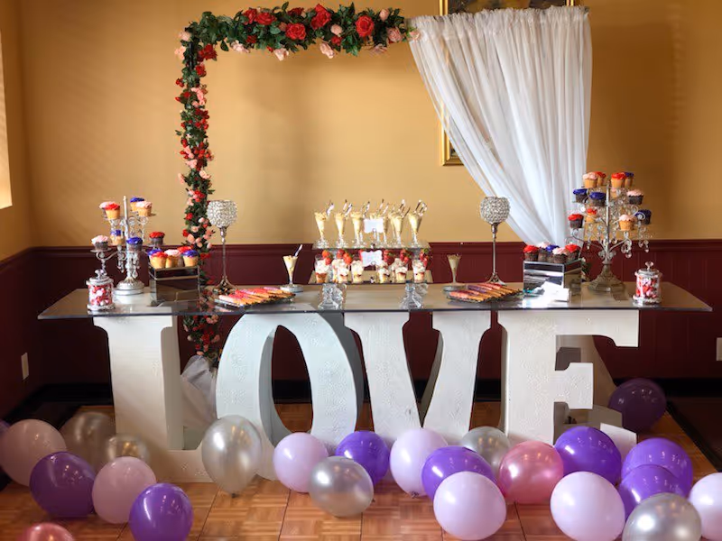 Decorated dessert table with large LOVE letters, arch, balloons, and white drapes in an indoor hall.