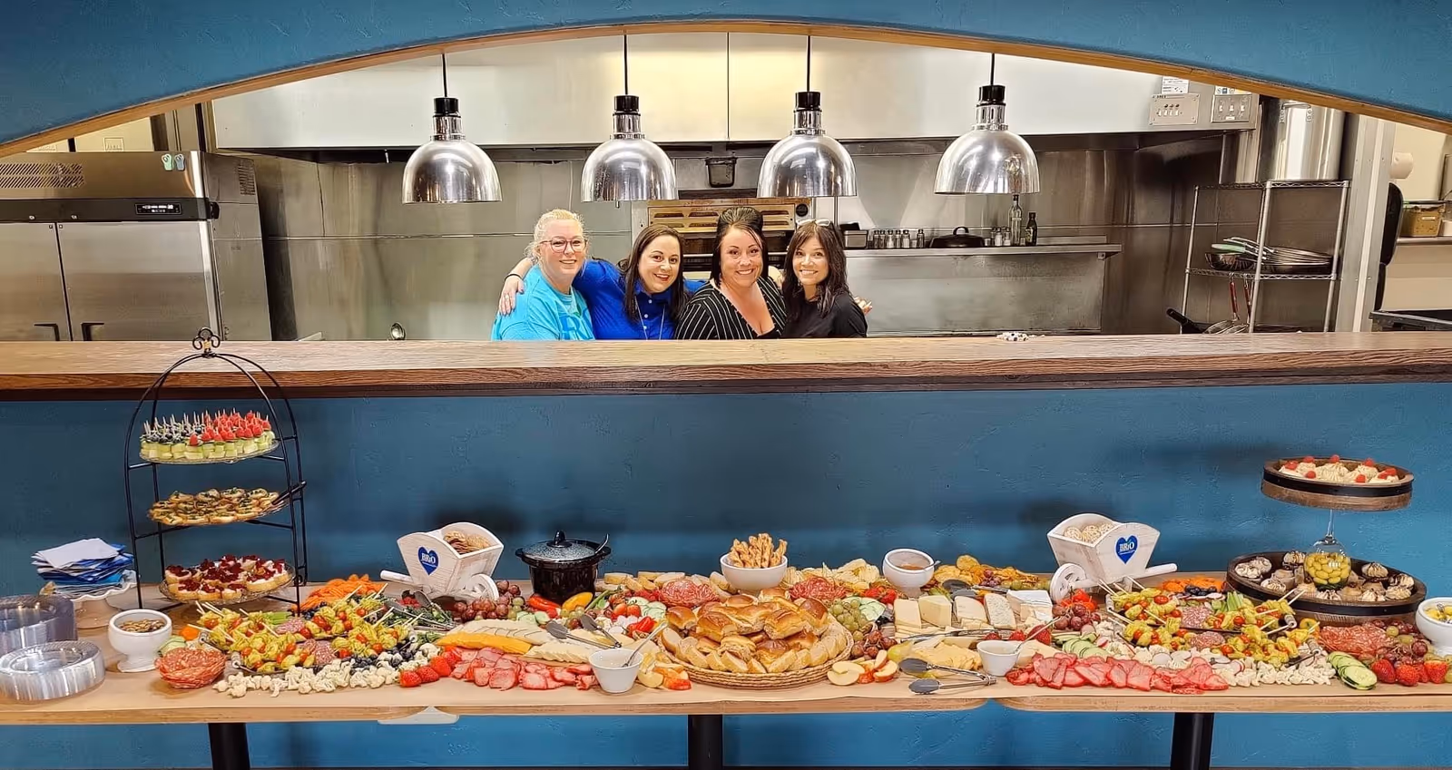 Long serving table laden with foods in a blue kitchen, four staff behind a pass-through.