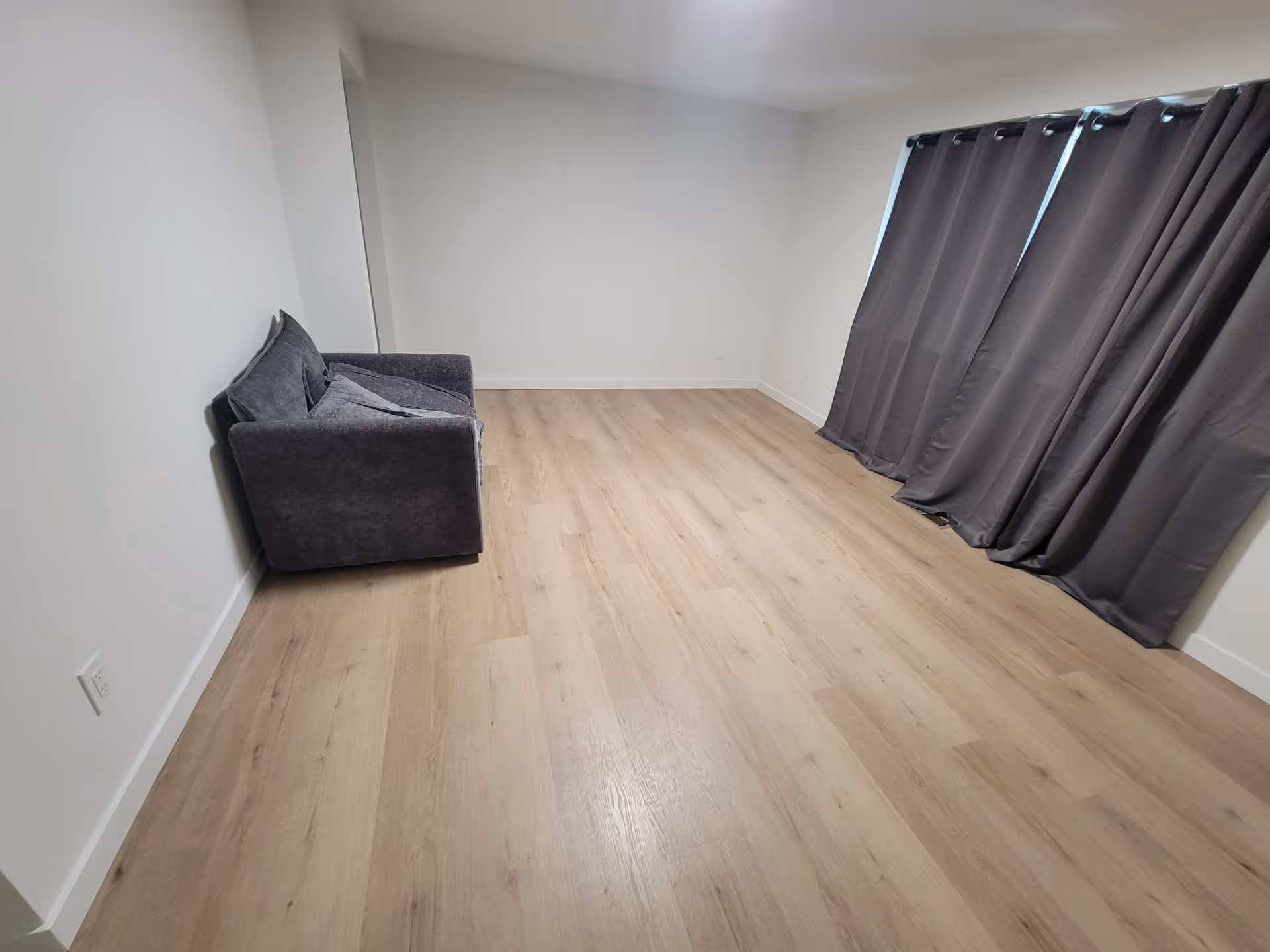 Minimal living room with gray sofa, light wood floor, white walls, and long gray curtain.