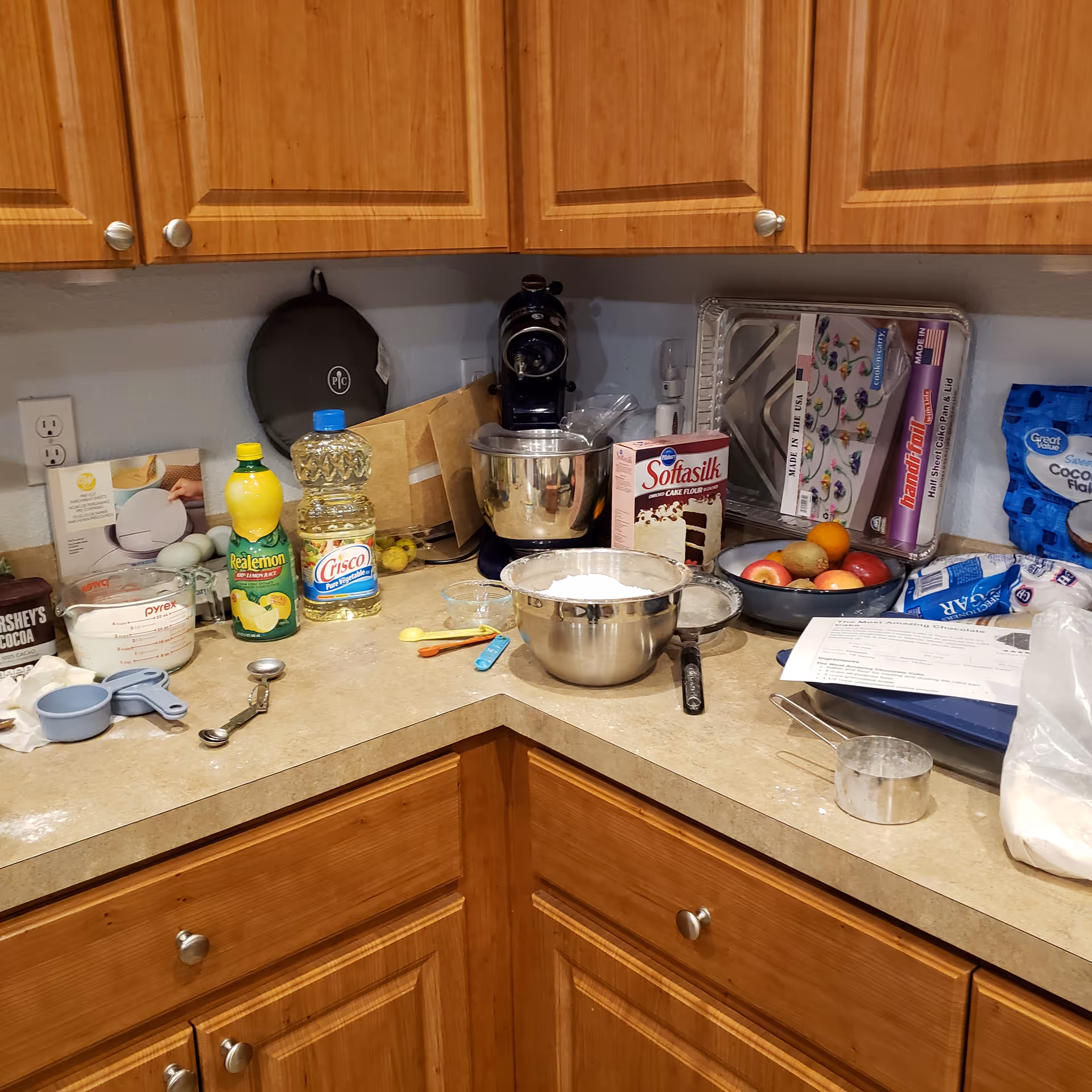 Cluttered kitchen counter with mixer, ingredients, and baking supplies under wooden cabinets.