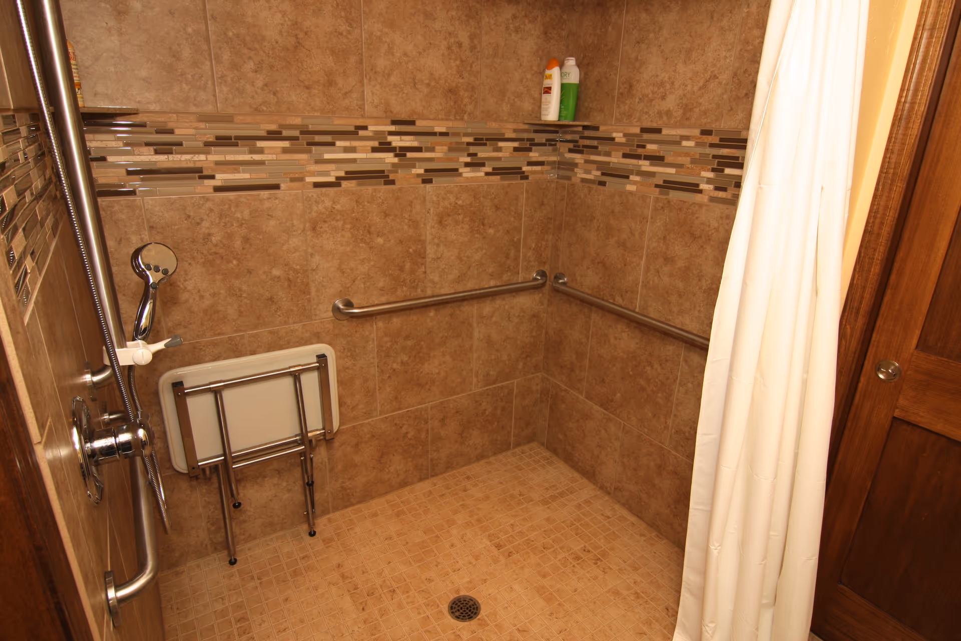 Accessible bathroom with tiled walls, grab bars, shower curtain, and a folding chair.