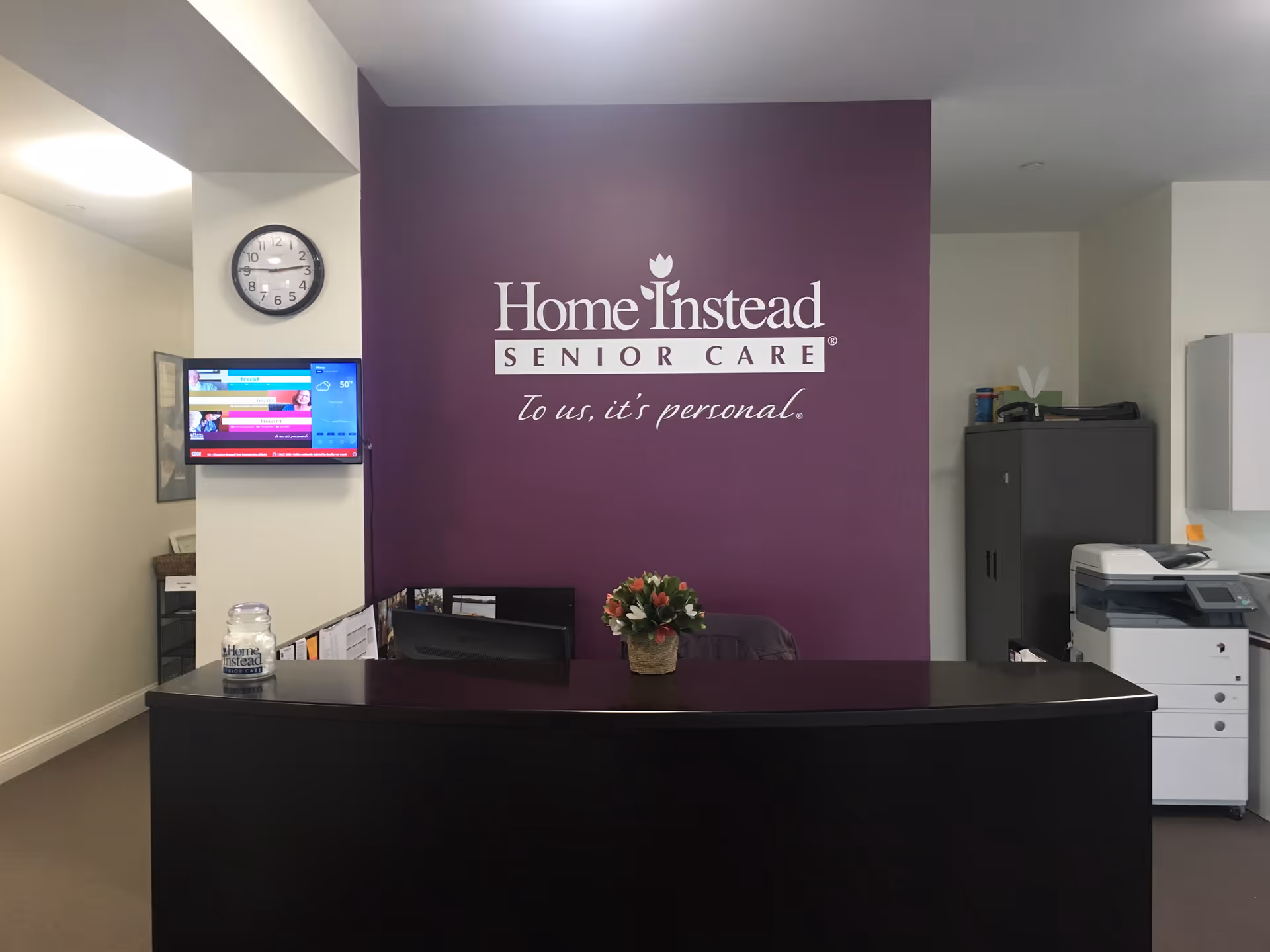 Reception desk in a Home Instead lobby with purple logo wall behind.