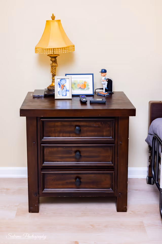 Dark wooden dresser with lamp, framed photos, and a figurine beside a bed.